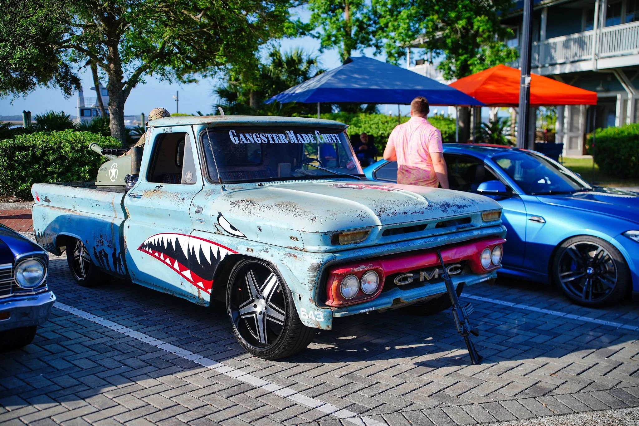 A vintage GMC pickup truck with a shark mouth and 'Gangster Made Chevy' written on the windshield, parked among modern cars at an outdoor event with tents and trees in the background.