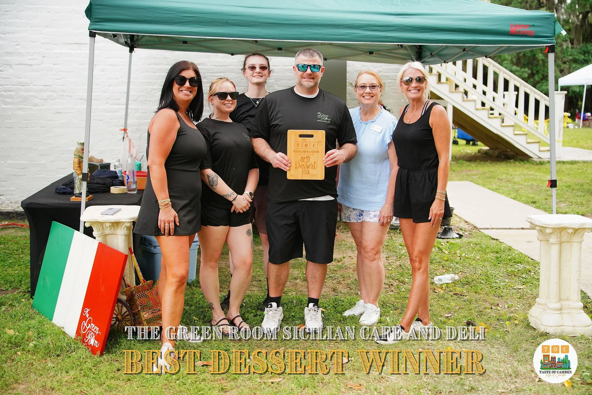 Group of six people standing outdoors under a green canopy, smiling, with a sign that reads 'Best Dessert 2026.' They are dressed in casual summer clothing, with some wearing sunglasses. A table with items and an Italian flag is visible on the left, 