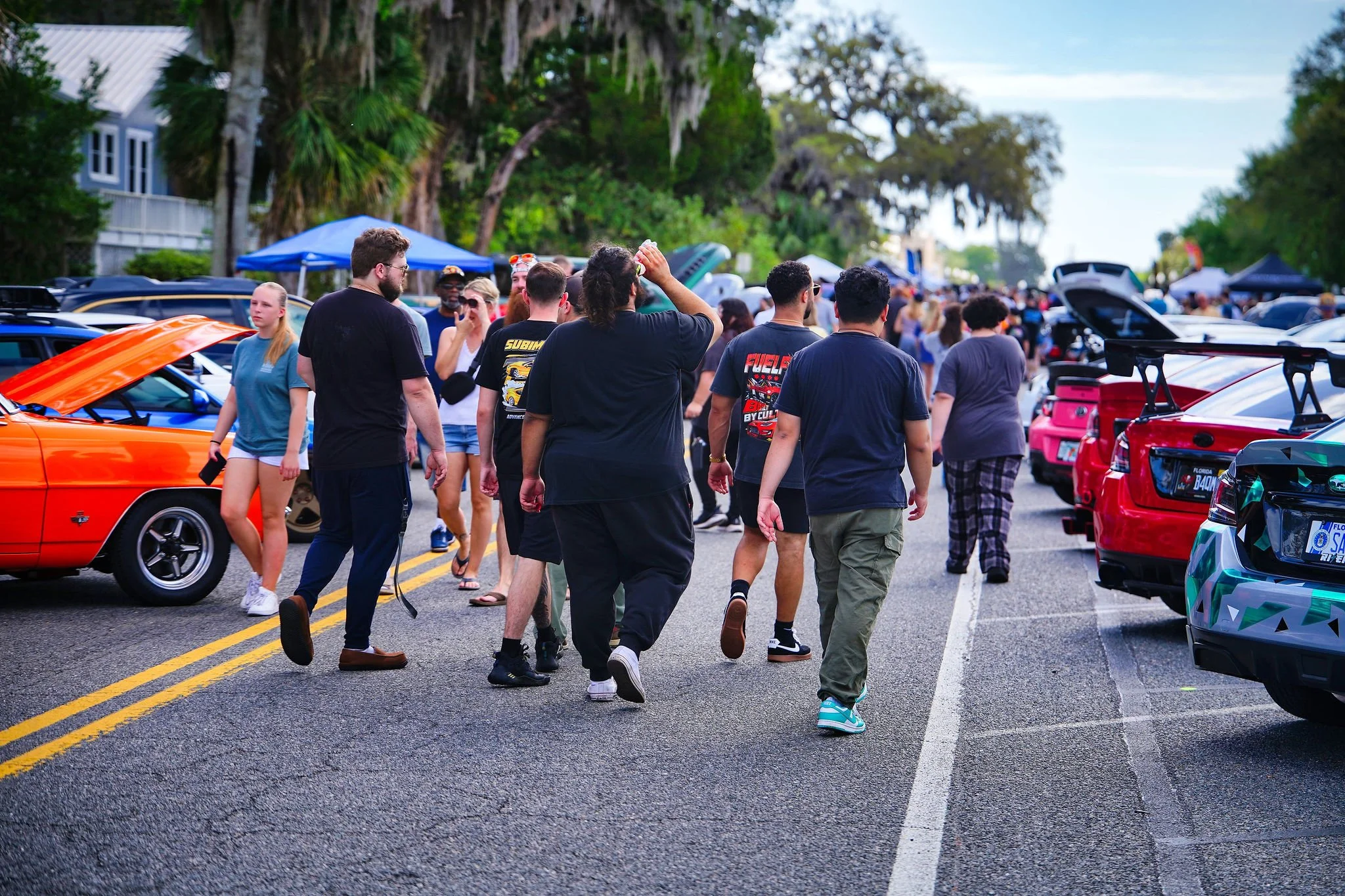 Crowd of people walking on a street lined with cars during a car meet or outdoor event with trees and blue sky in the background.