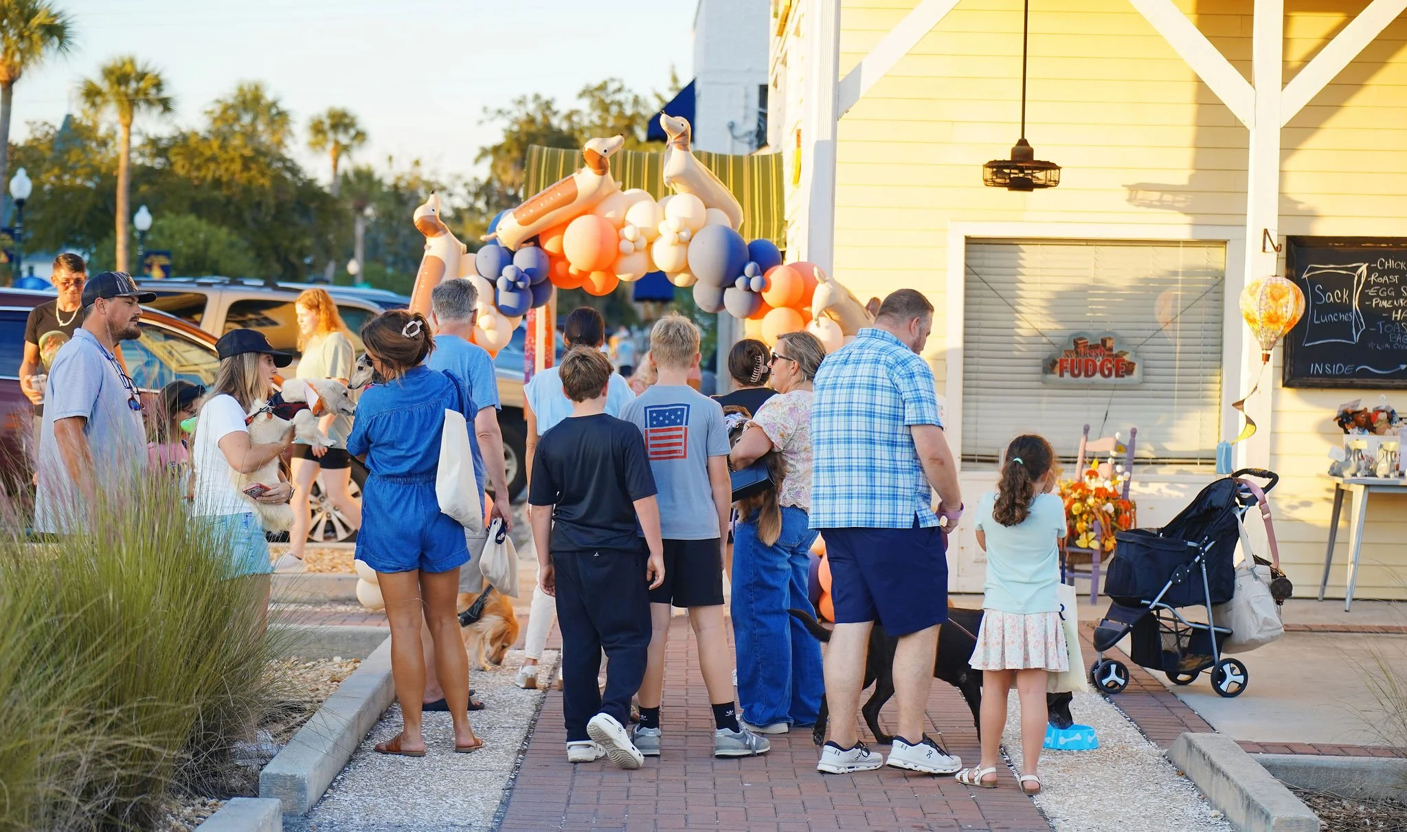 People gathered outside a yellow building with balloon decorations, outdoor signage, and children with their guardians, during an outdoor event or festival in the evening.