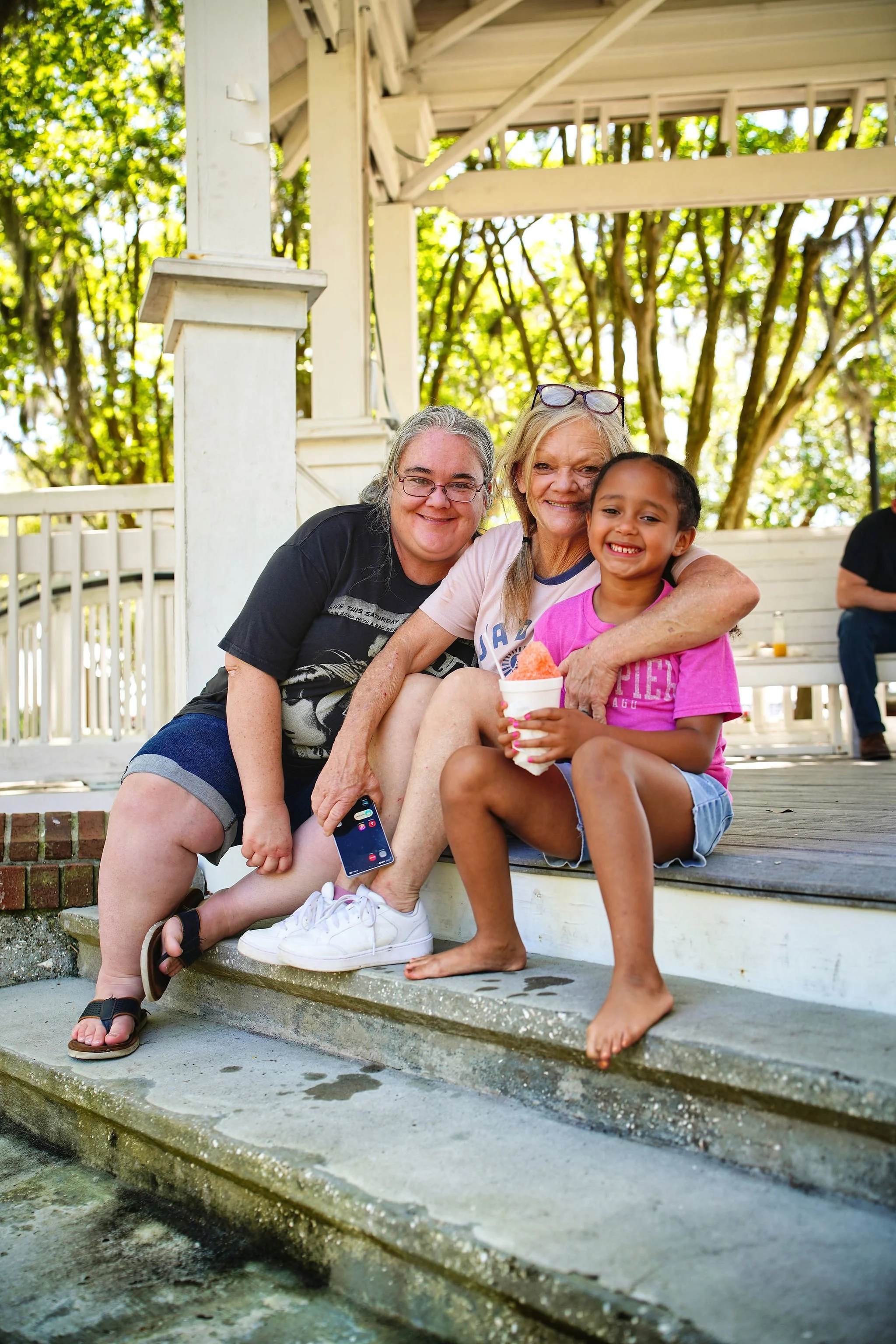 Three women and a girl sitting on a porch step, smiling at the camera, with trees in the background. The girl holds a cup of shaved ice.