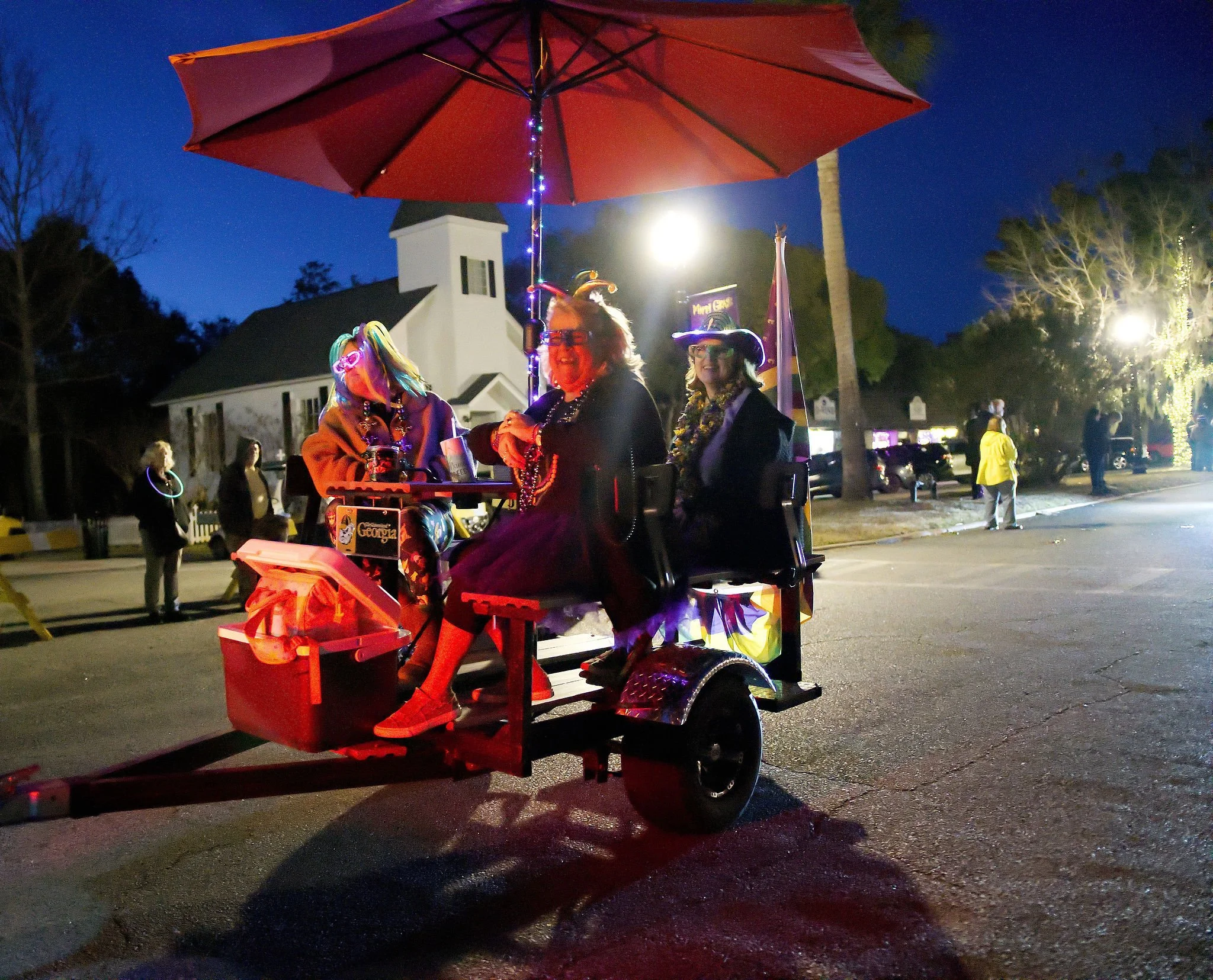 Nighttime parade scene with women dressed in costumes riding a decorated cart with an umbrella, illuminated by streetlights and camera flash, and a church and trees in the background.