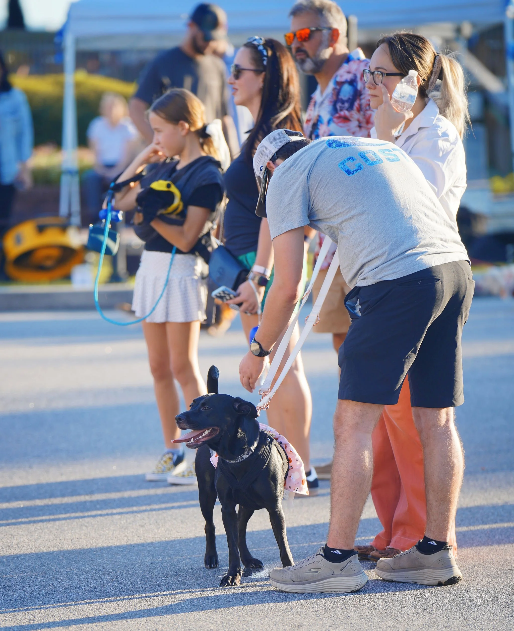 People standing outdoors on a sunny day, with a black dog in the foreground, and a waterfront or marina in the background.