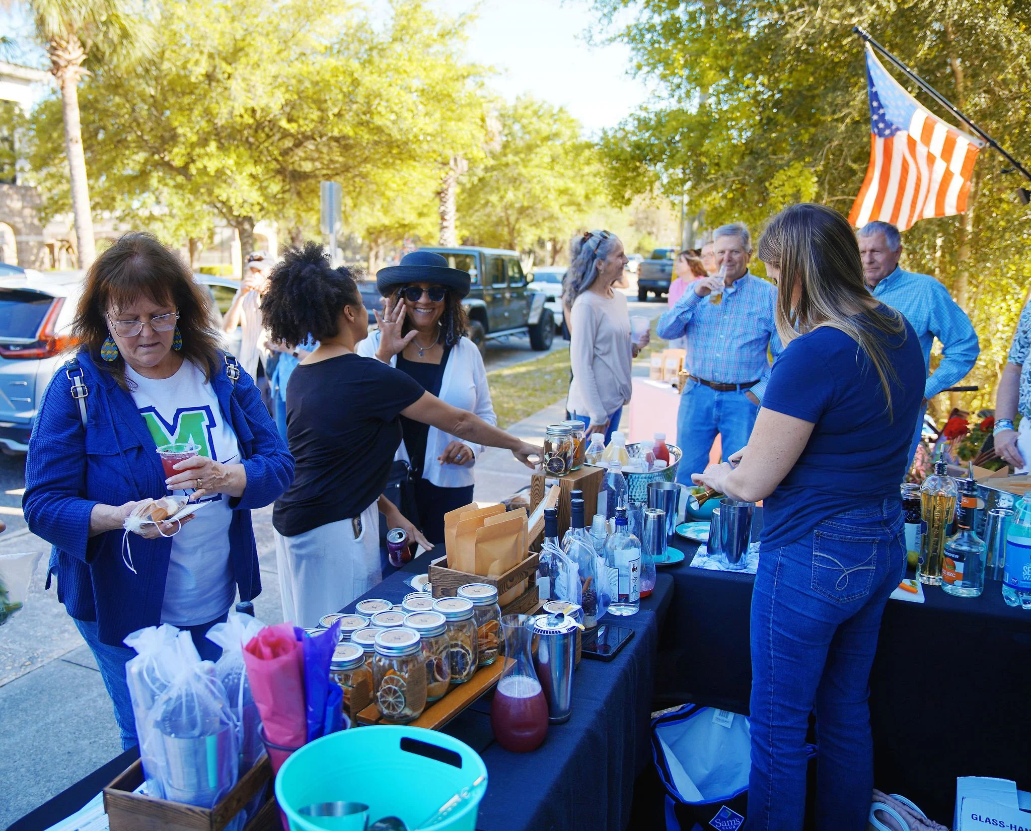 People shopping at an outdoor market stand with beverages and jars, with American flags in the background and trees surrounding the area.