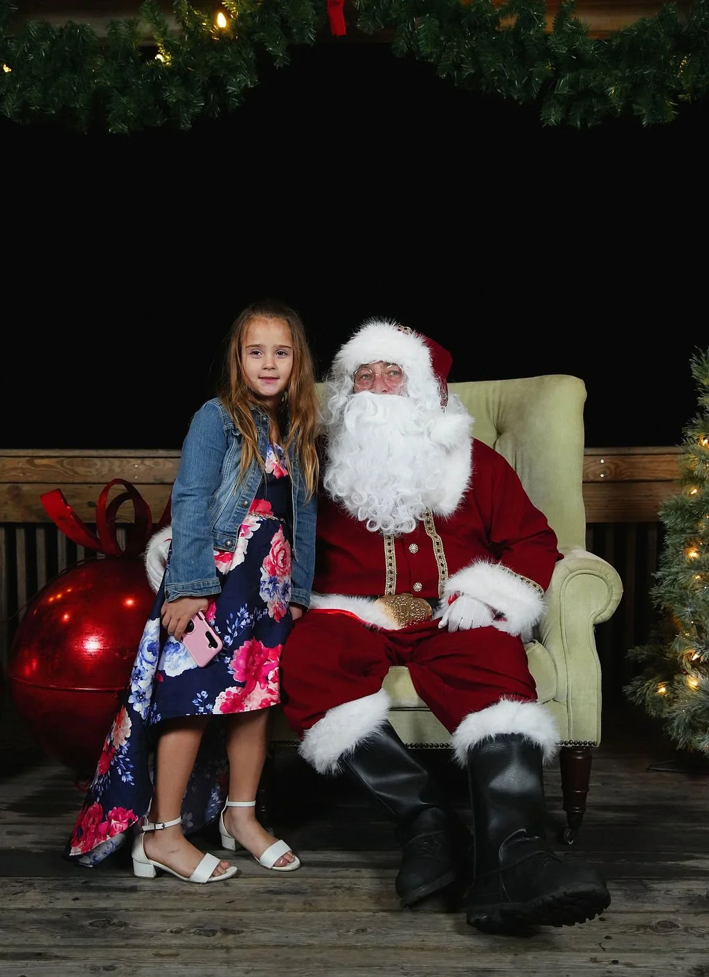 A young girl standing next to Santa Claus, who is sitting in a green armchair, during a Christmas event, surrounded by Christmas decorations including a large red ornament and a decorated Christmas tree.