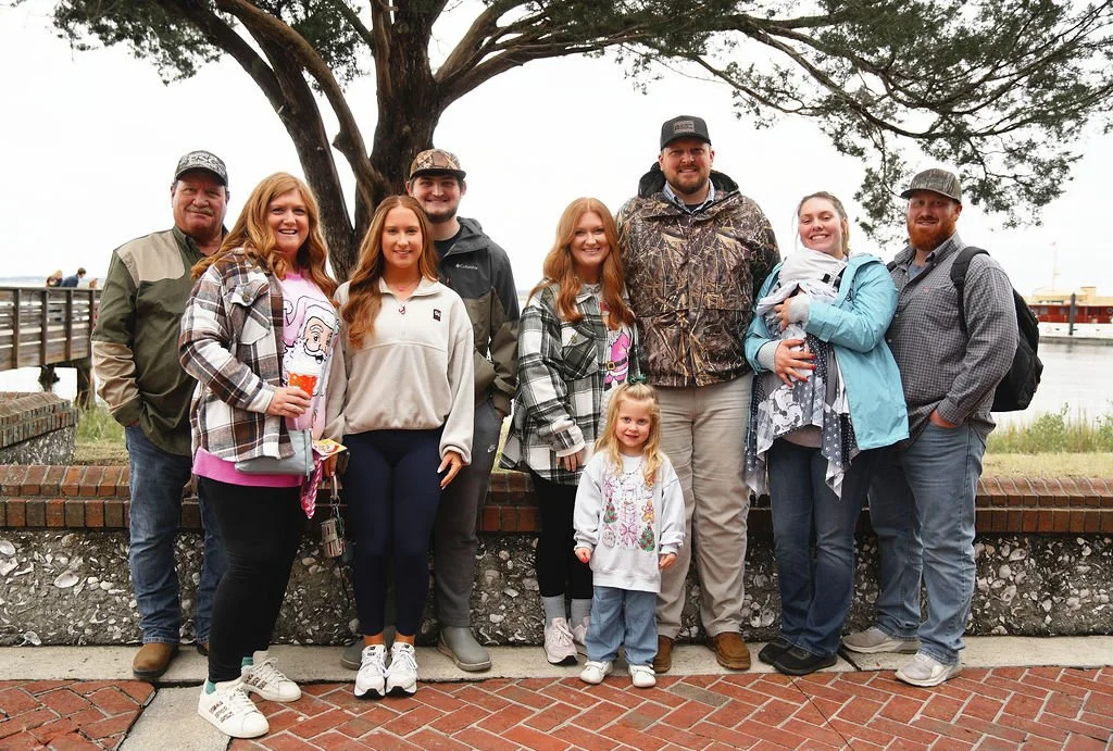 Family group photo outdoors in front of a large tree, with a body of water and boats in the background. The family includes adults and children dressed in casual, slightly cooler weather clothing.
