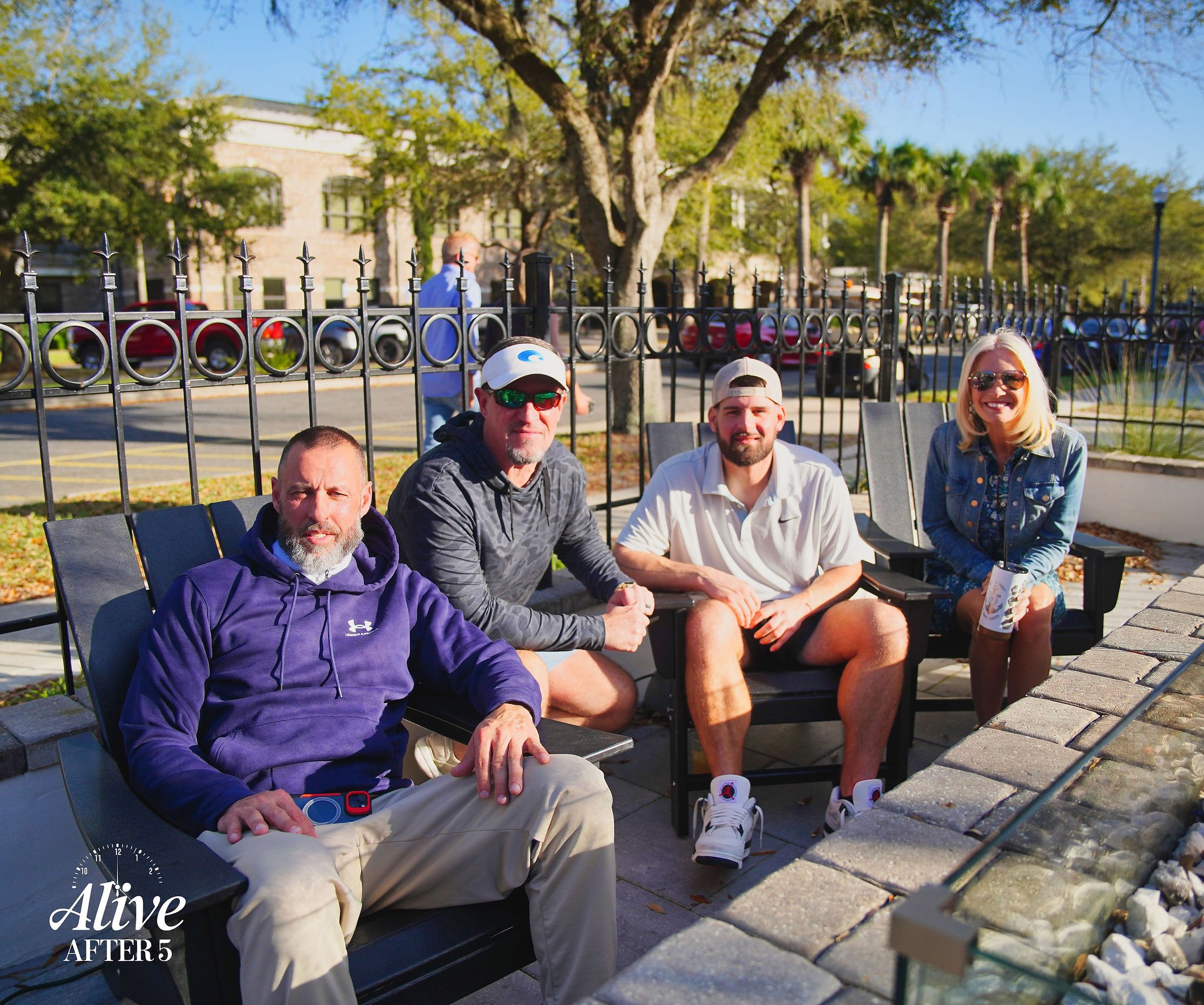 Four people sitting on outdoor benches in a park, smiling at the camera, with trees and parked cars in the background.