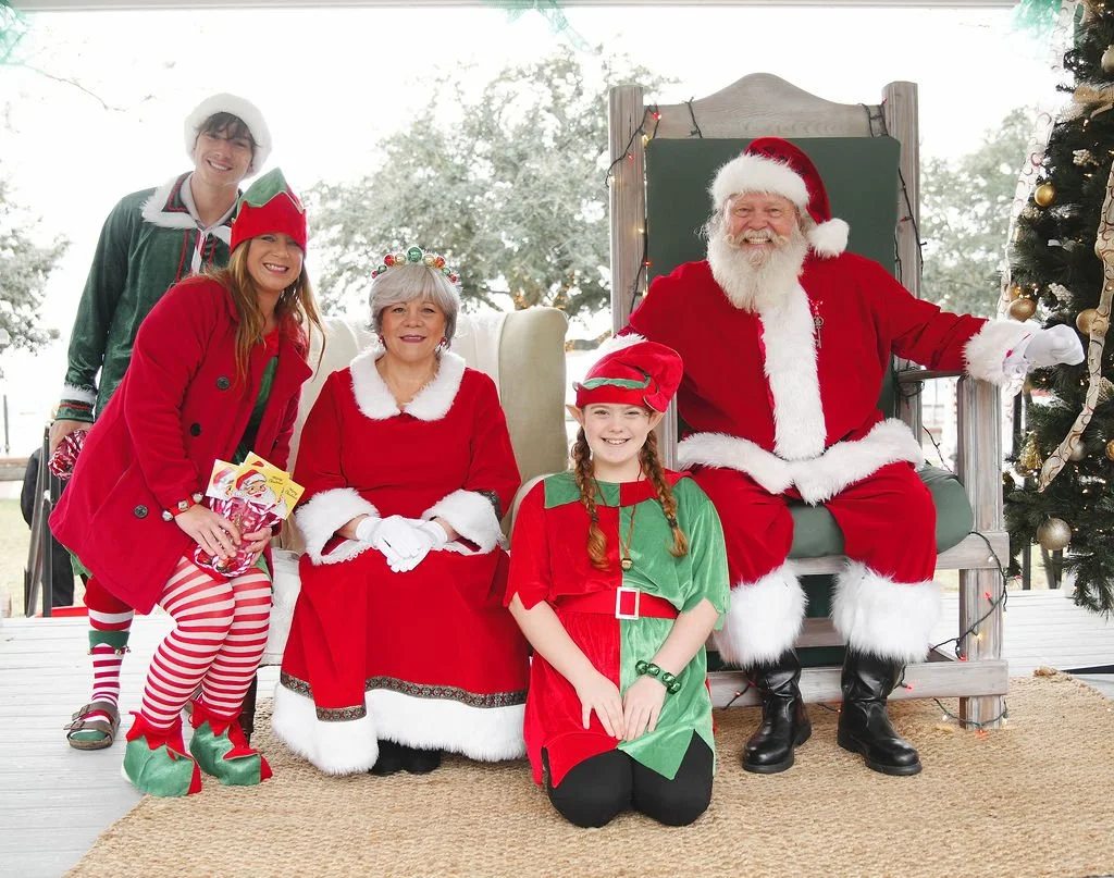 Group of people dressed in Christmas costumes with Santa Claus, elves, and Mrs. Claus, sitting and standing in front of a decorated Christmas tree.