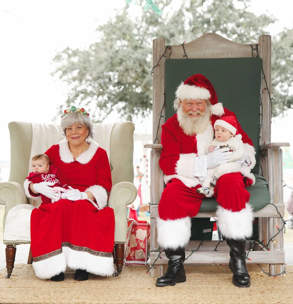 Santa Claus and Mrs. Claus sitting with two babies, one on each person's lap, during a Christmas event. Mrs. Claus is sitting on a white armchair, and Santa is seated on a wooden throne with Christmas lights.