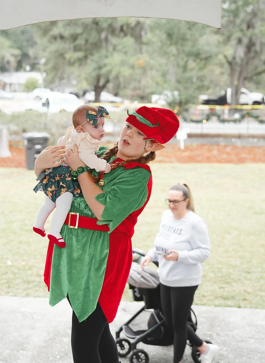 A woman dressed as an elf holding a baby girl during a festive outdoor event. The woman is wearing a red and green elf costume with a matching hat, and the baby is wearing a headband with a bow, a cream top, and a skirt with holiday print. A woman wi
