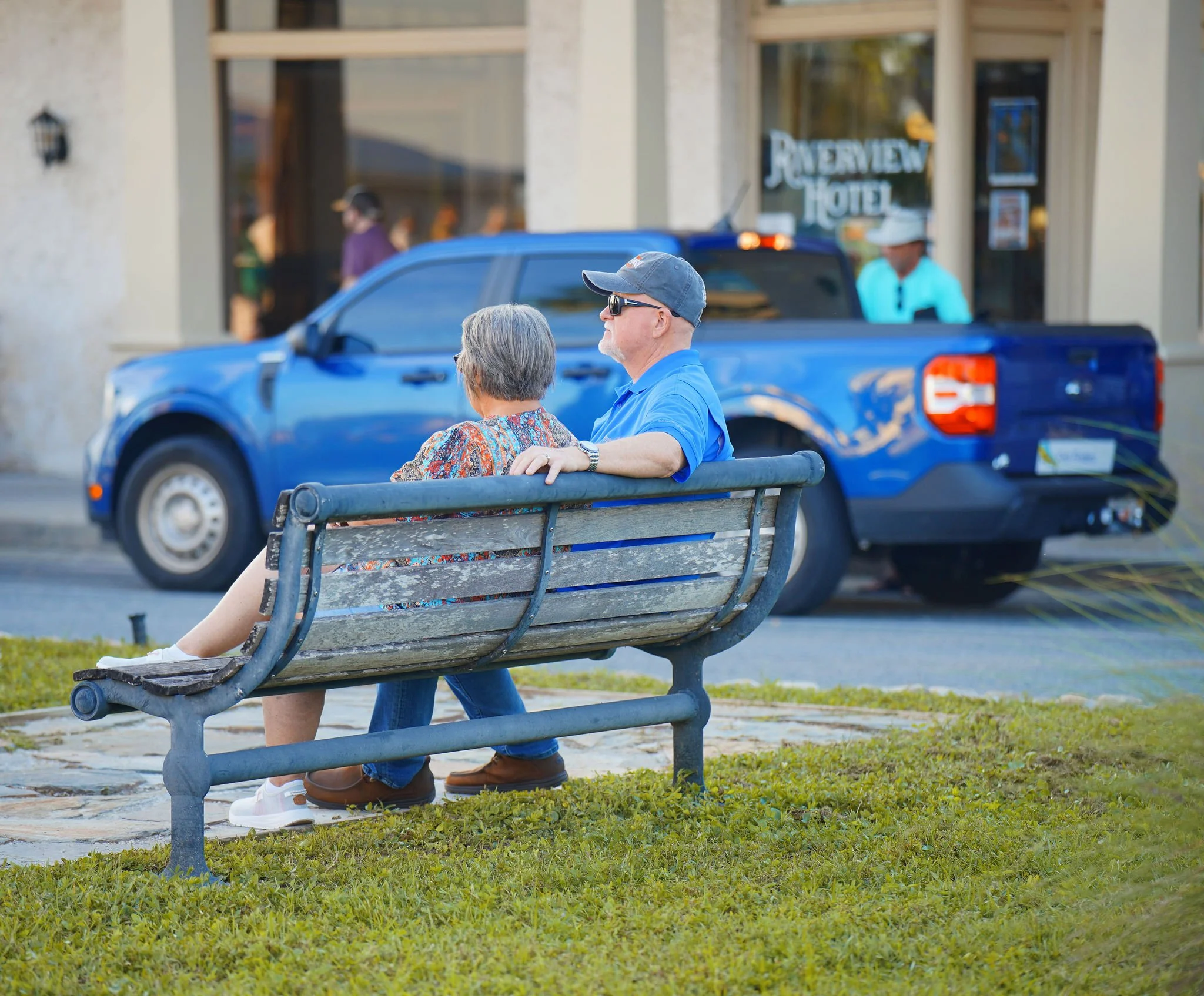 A man and woman sitting on a public park bench, with the man leaning in towards the woman, who has gray hair, and the man is wearing sunglasses, a gray cap, and a blue shirt. They are inside a city area with a blue pickup truck parked nearby and a ho
