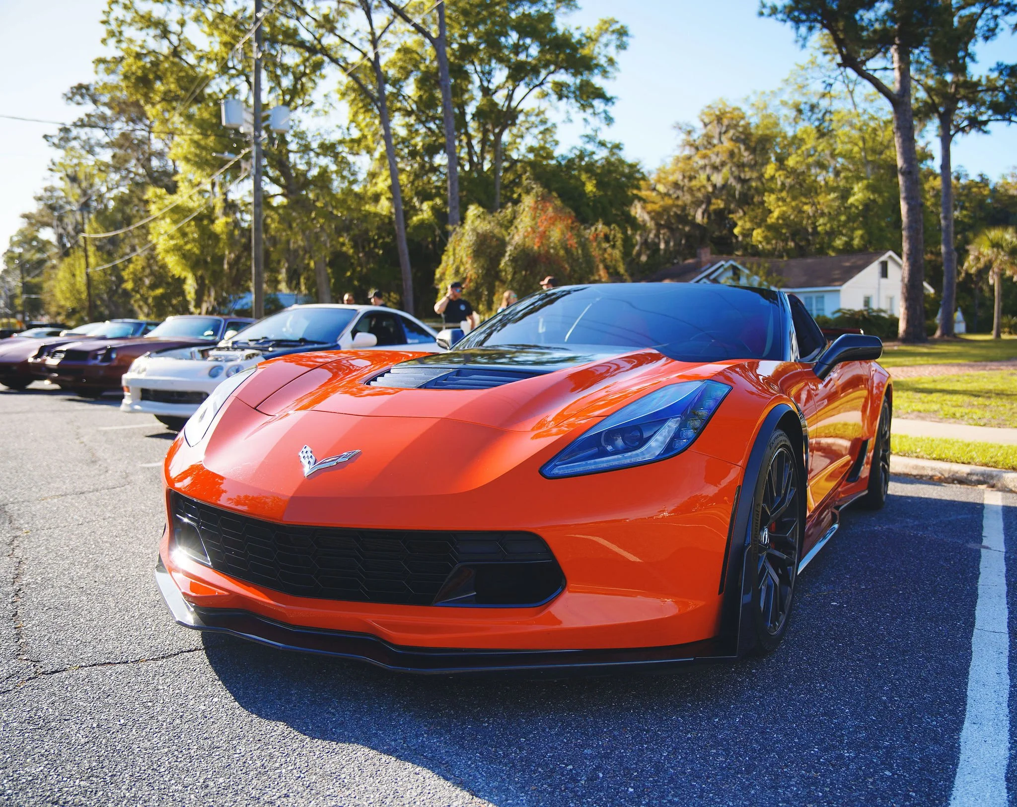Orange Corvette sports car parked at a car show with other vehicles and trees in the background.