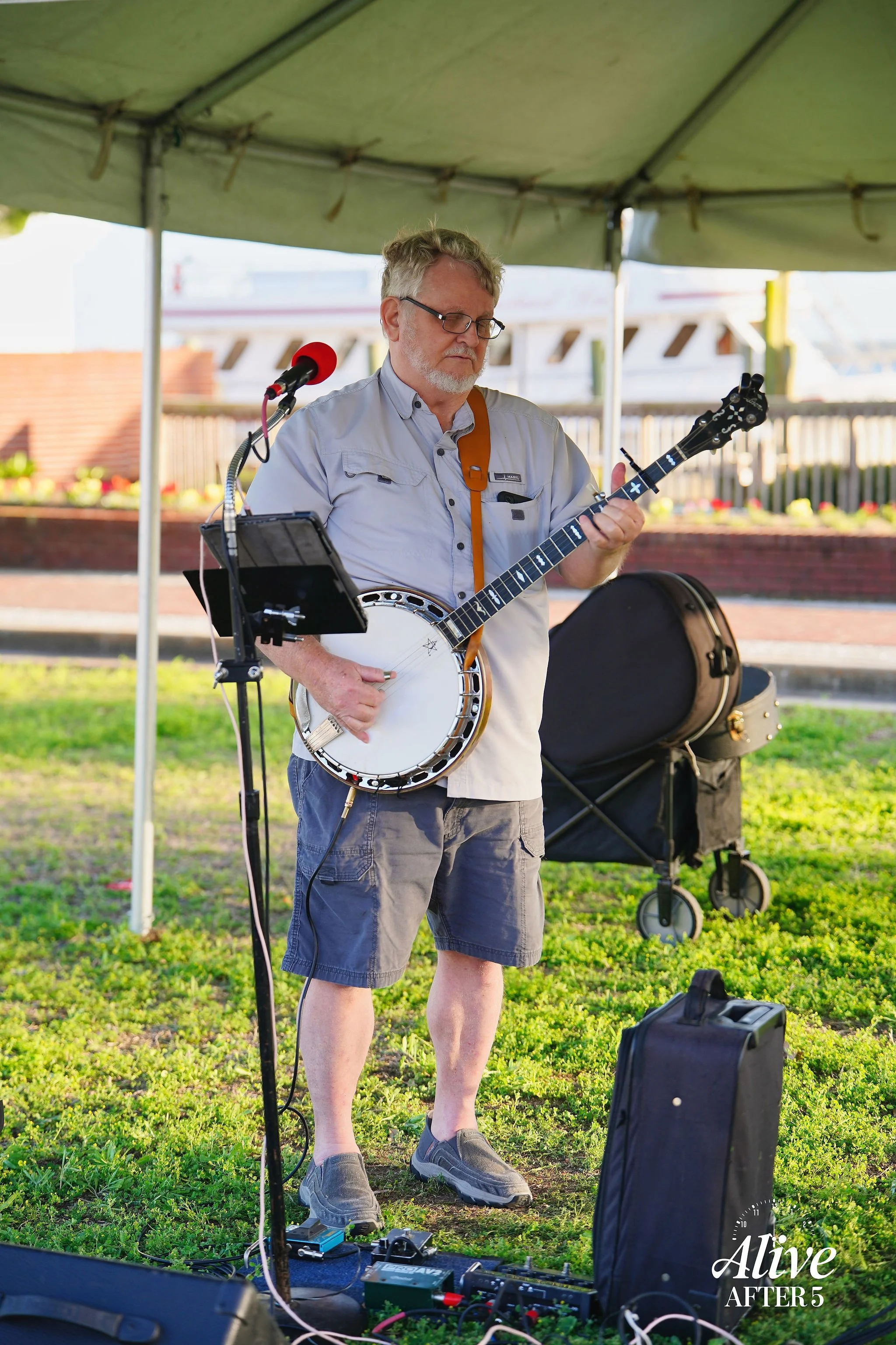Man playing banjo and guitar under a green tent, with audio equipment on the grass in front of him, and a stroller nearby, outdoors during daytime.