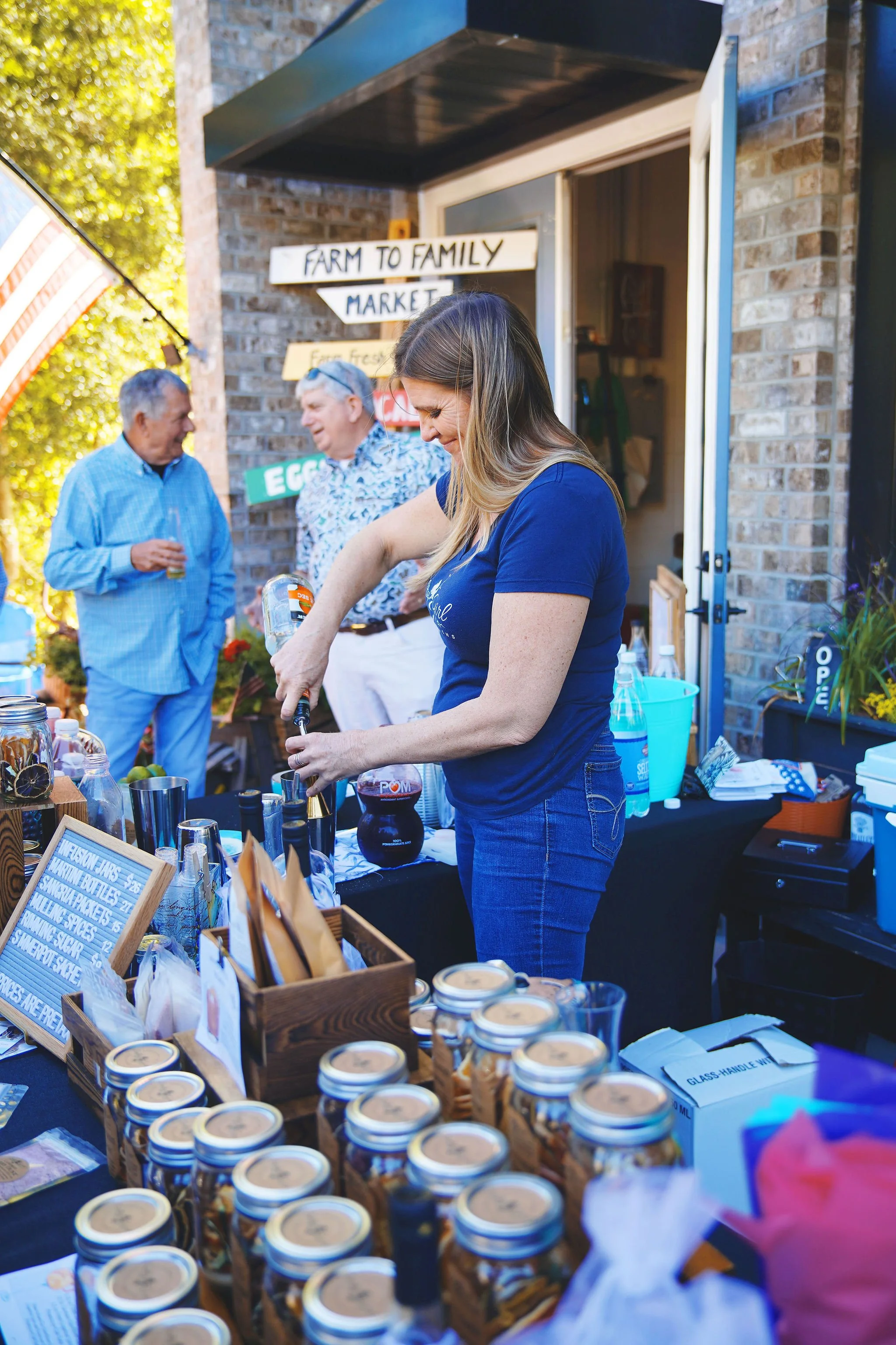 Woman preparing drinks at a farm-to-family market stall with jars, jars, and a chalkboard sign in the foreground, and two men talking in the background near a brick building.