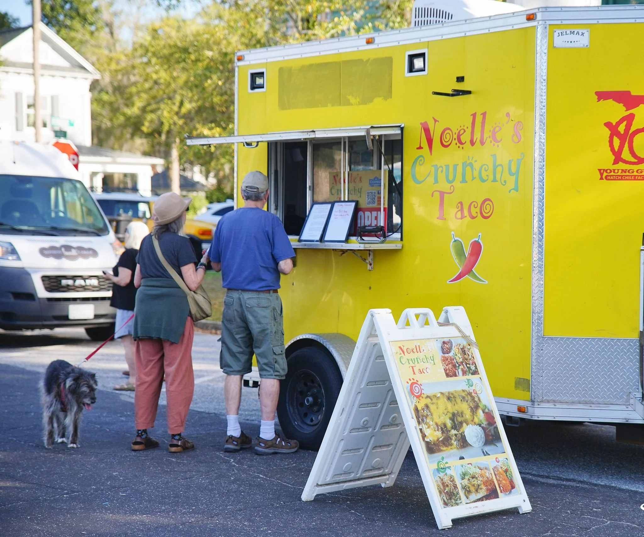 Yellow food truck named 'Noelle's Crunchy Taco' with a menu board outside, serving tacos. Two customers, one with a dog on a leash, are placing orders at the window. There are cars in the background and trees with green leaves.