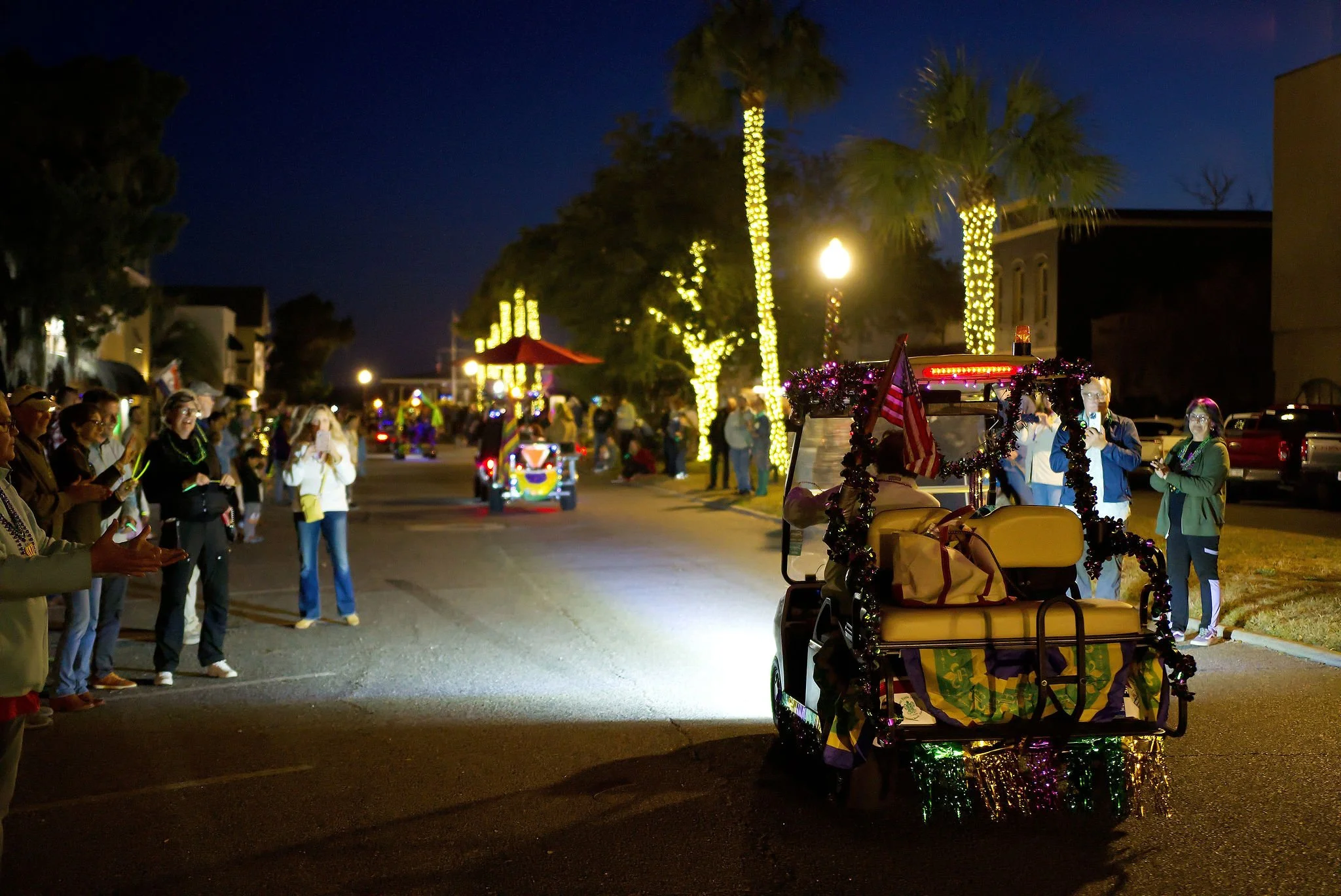 Nighttime parade with decorated vehicles, lined by spectators taking photos, with trees wrapped in string lights and illuminated buildings in the background.