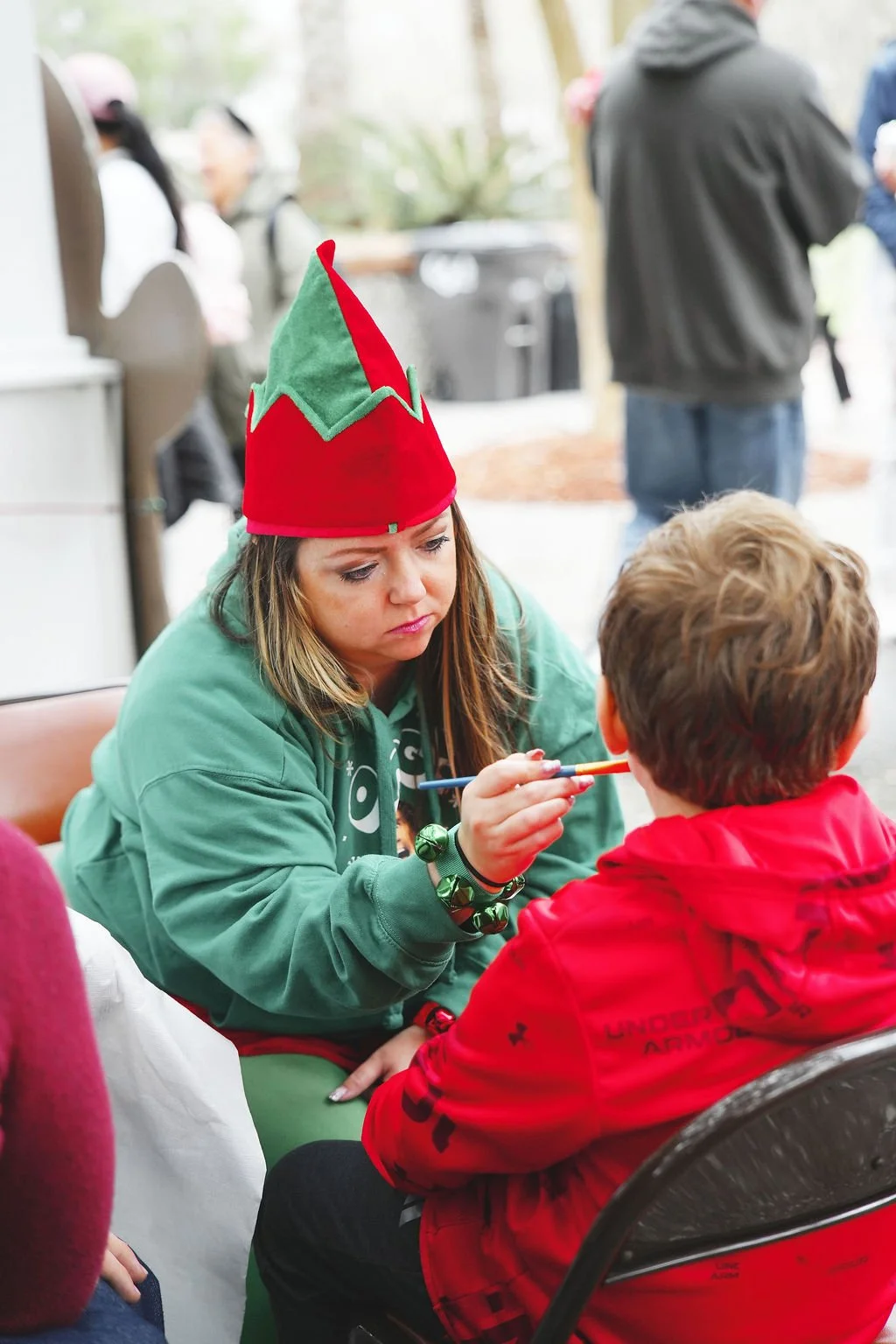 A woman dressed as an elf is painting the face of a young boy at a holiday event. She is wearing a red and green elf hat and a green sweatshirt.
