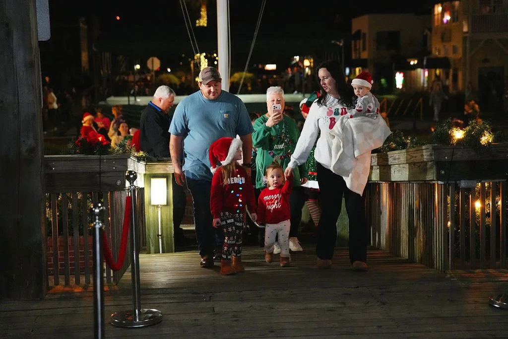 Family walking on a decorated Christmas pier at night, with Christmas lights and people in festive attire, some wearing Santa hats.