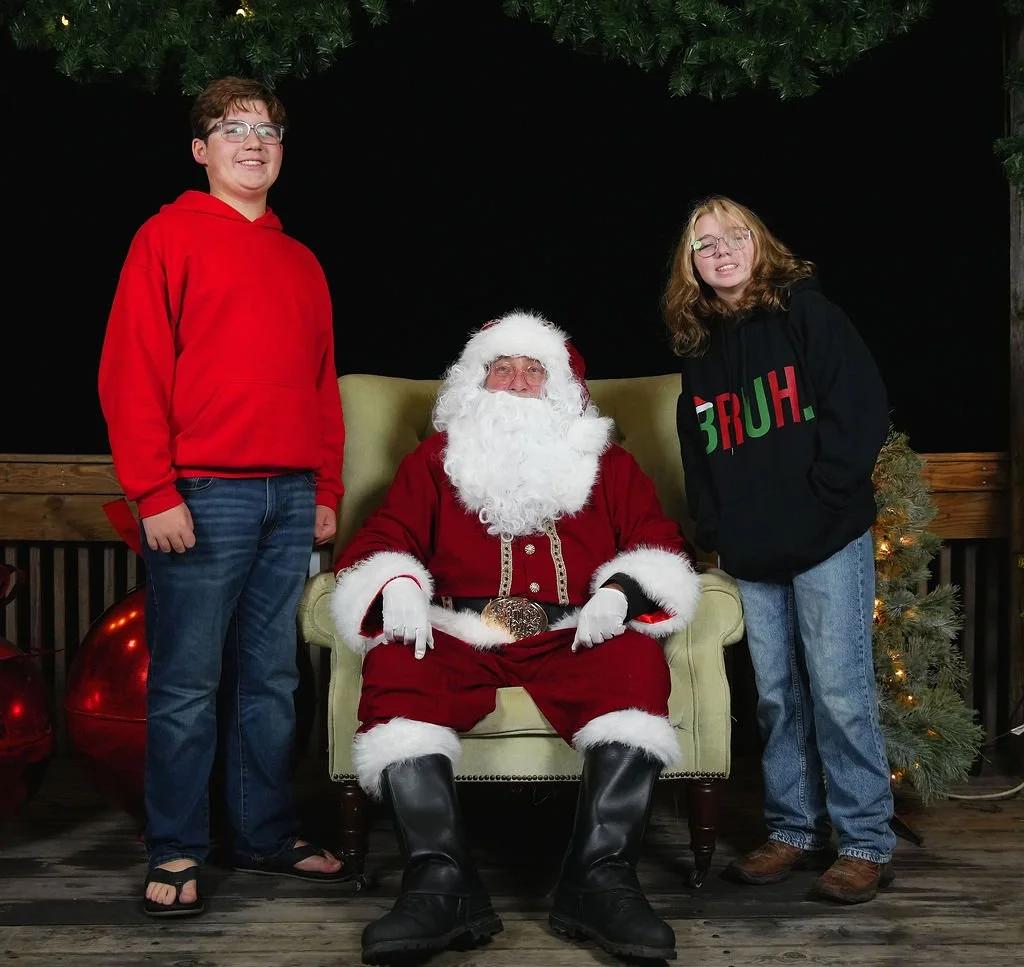 Two children, a boy in a red hoodie and a girl in a black hoodie with 'BRUH.' printed on it, standing next to a man dressed as Santa Claus sitting on a chair. The scene is decorated with Christmas trees and ornaments.