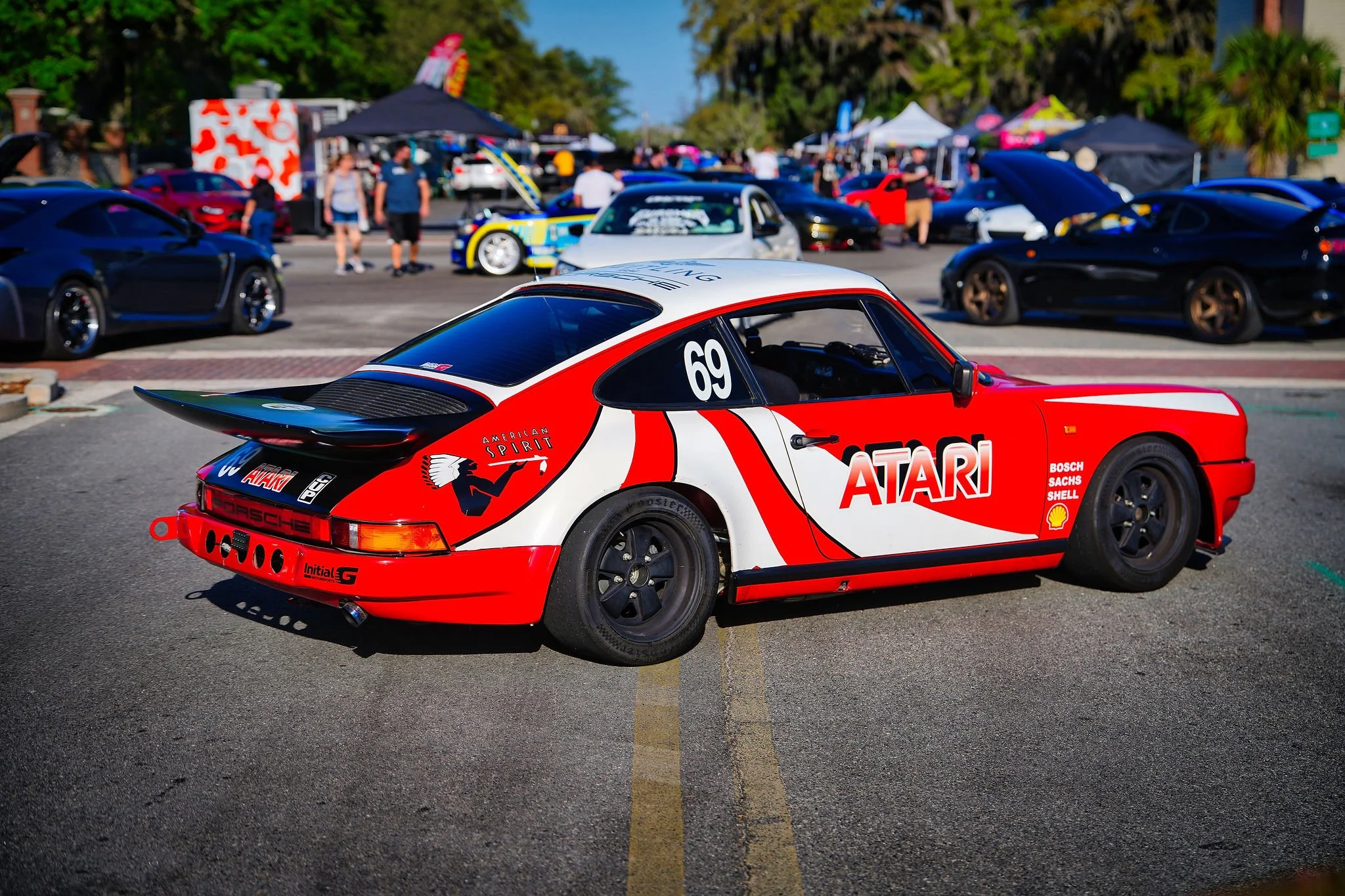 Red and white vintage Porsche race car with large rear spoiler, number 69, and various sponsor logos, parked at a car show with other sports cars and tents in the background.