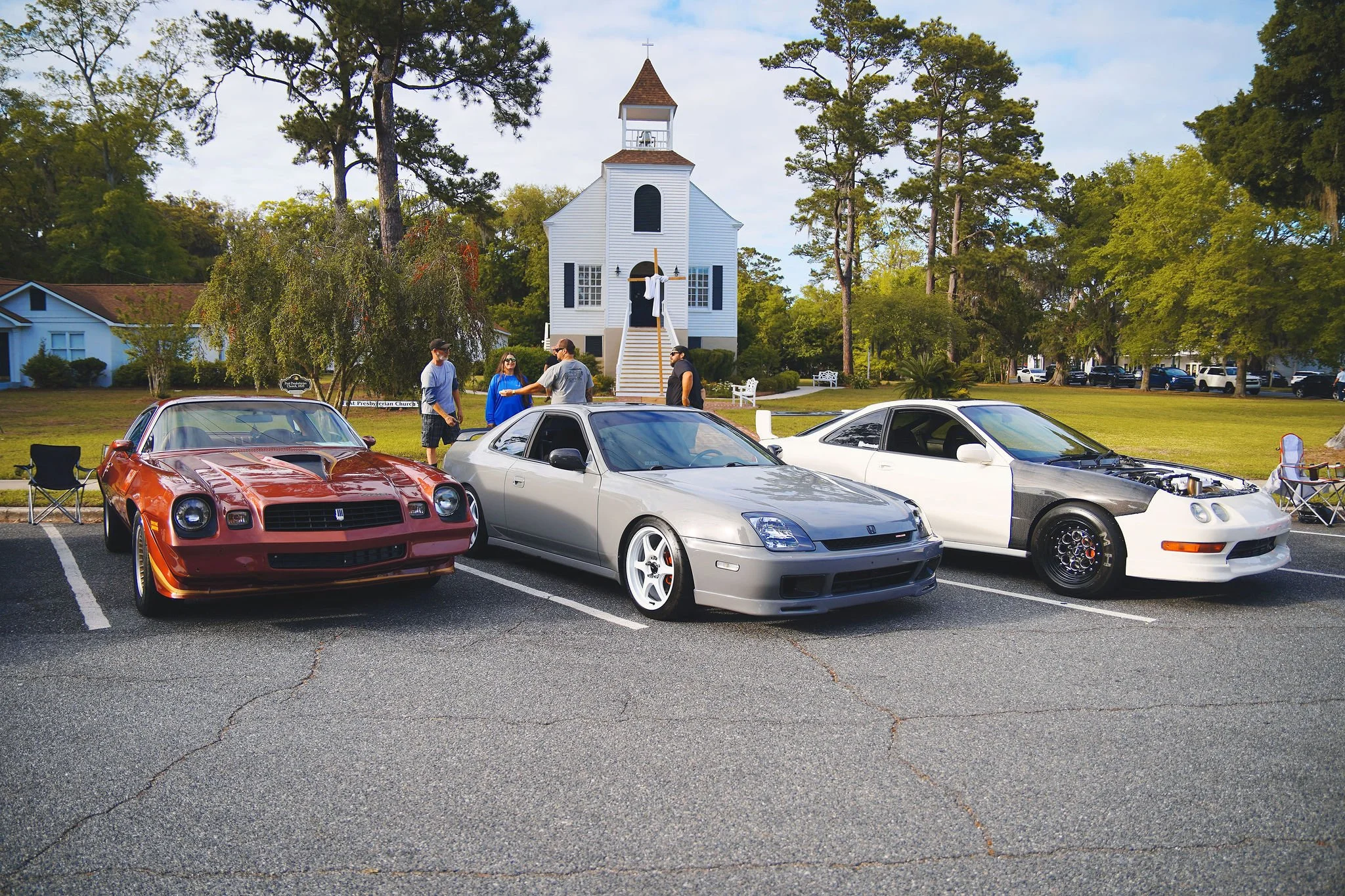 Three classic sports cars parked in a lot in front of a white church with a bell tower and wooden steps. People are talking and walking near the cars and church, with trees and a grassy area in the background.