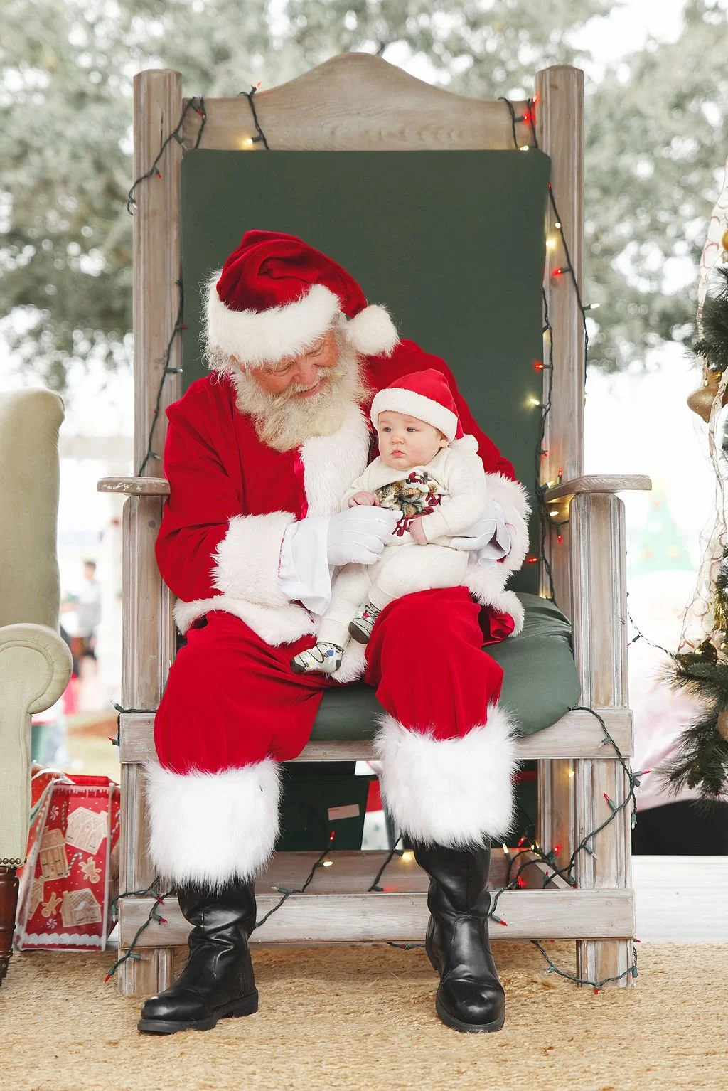 Santa Claus dressed in a red suit with white fur trim, sitting on a wooden throne with greenery and Christmas lights, holding a young child in Christmas attire.