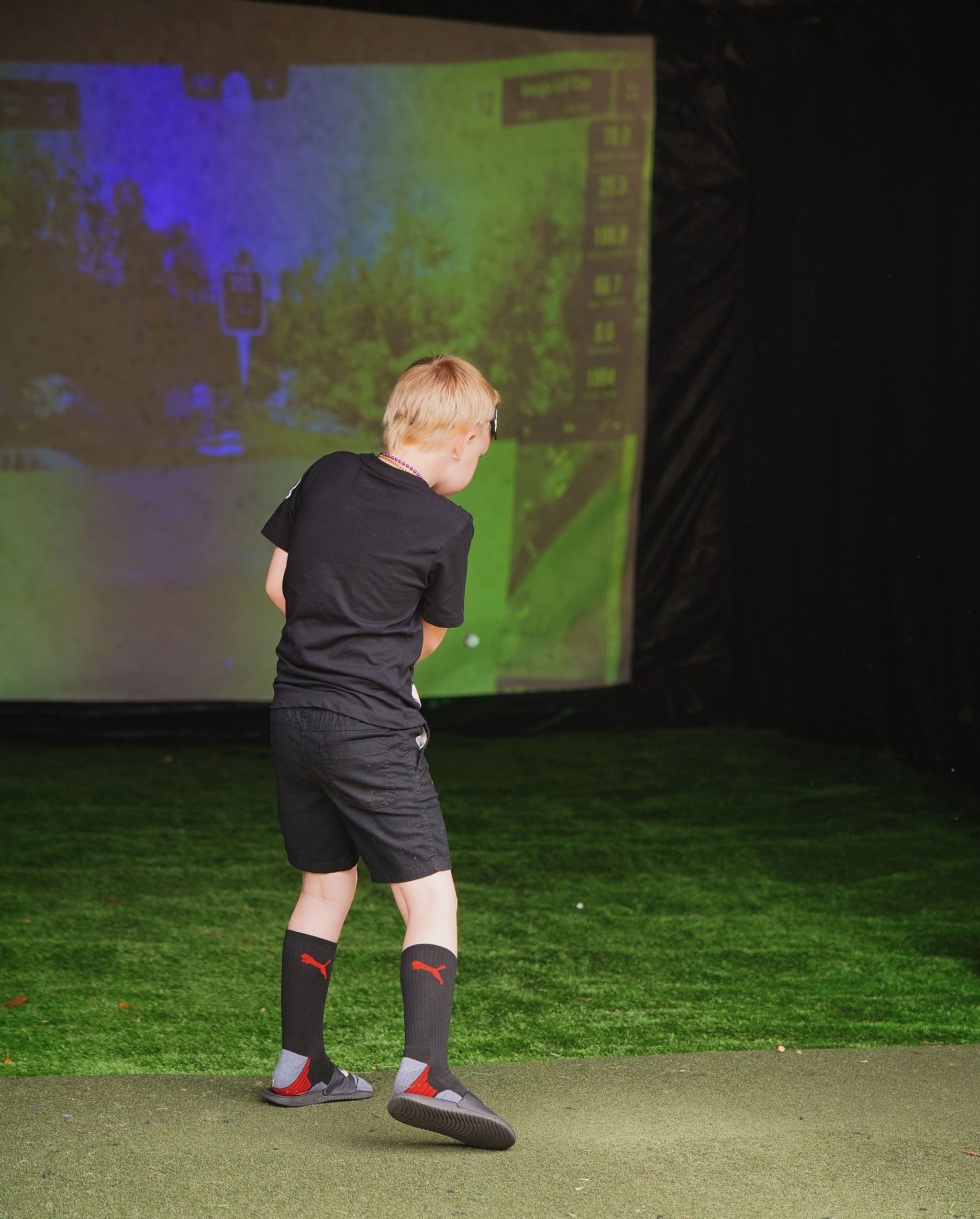 Young boy in black shirt, shorts, and Puma socks playing indoor golf, with a screen displaying a virtual golf course in the background.