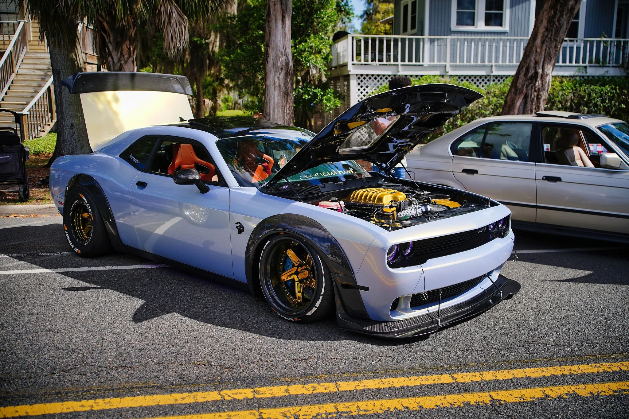 A modified sports car with its hood open parked on a street next to another car. The sports car has black accent wheels, a carbon fiber front splitter, and a yellow engine cover. It is silver with a black rear wing and orange interior seats.