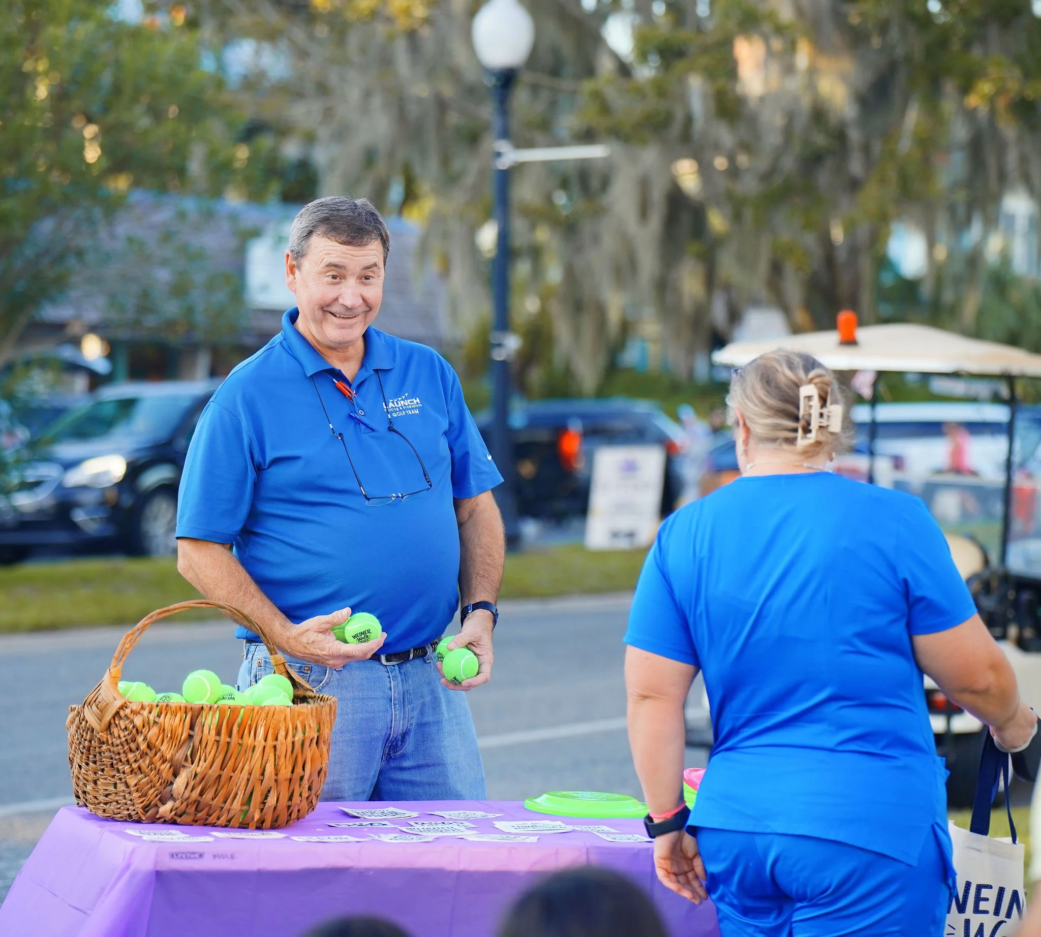 A man in a blue shirt with a purple table in front of him holding a basket filled with tennis balls, talking to a woman in a blue shirt during an outdoor event in the evening with trees and streetlights in the background.