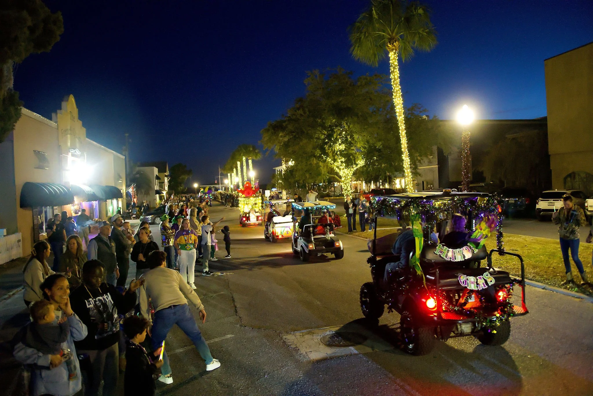 Nighttime parade on a street decorated with Christmas lights, featuring decorated golf carts and a crowd of people, some taking photos and watching the float procession.