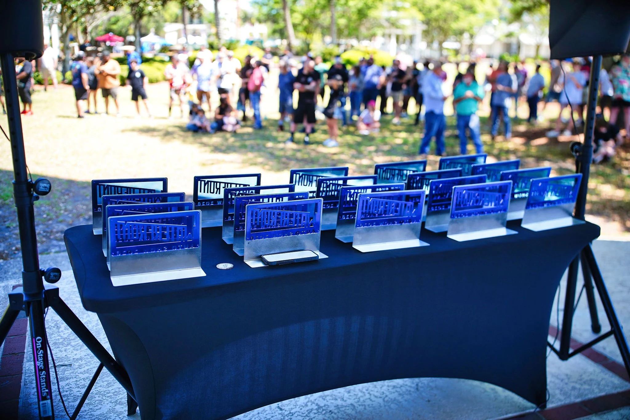 A table with several awards or trophies on it, set up outdoors with people gathered in the background at an event or festival.
