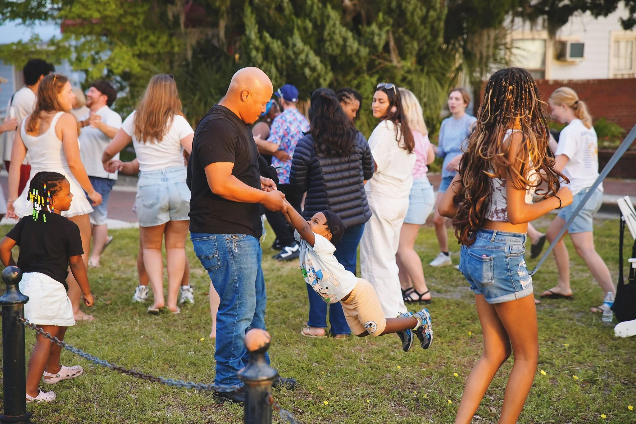 A group of children and adults at an outdoor party, with a young boy jumping into a man's arms. People are dancing and socializing on a grassy area with trees and houses in the background.