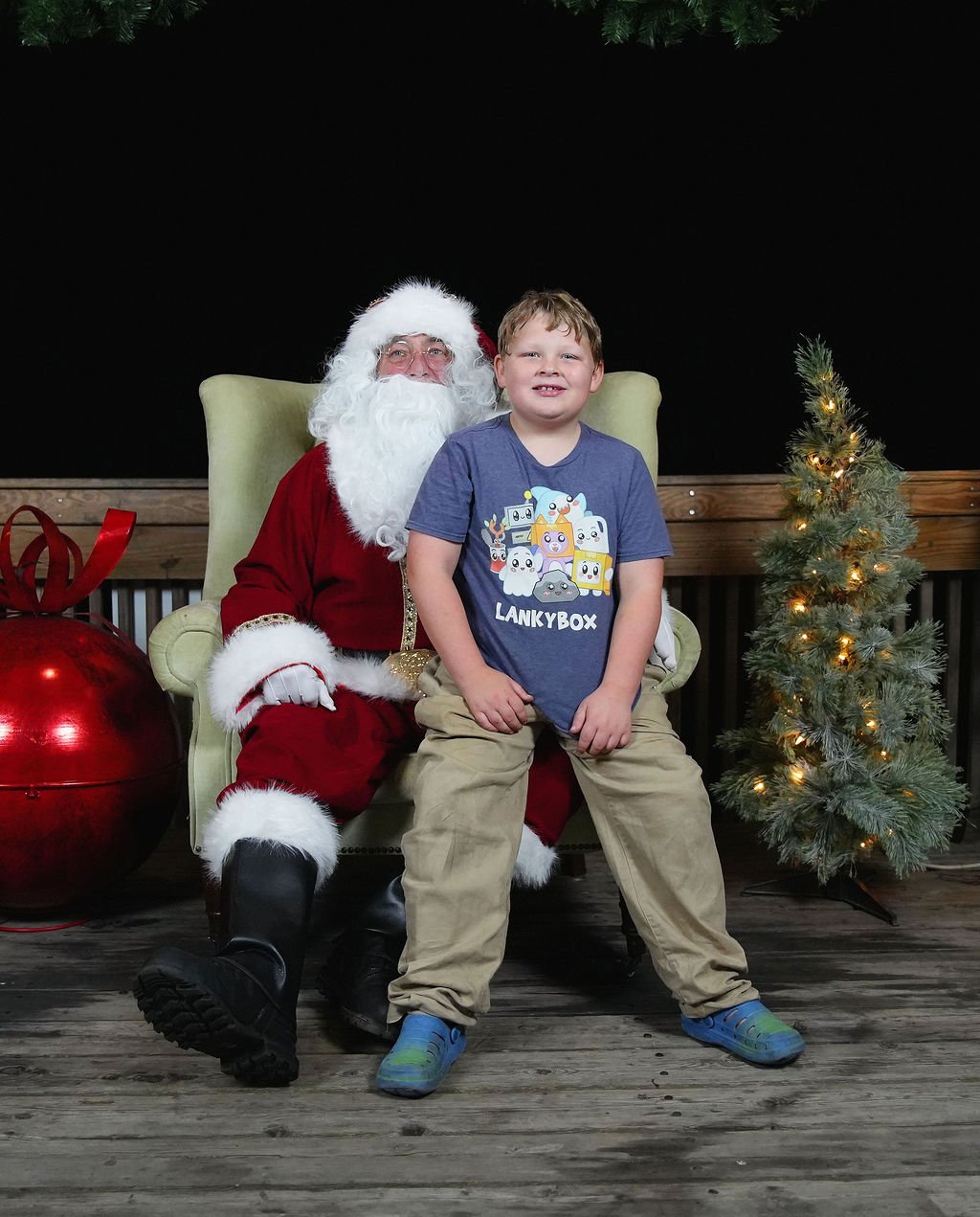 A boy sitting on Santa Claus's lap during Christmas. Santa is dressed in red and white, with a long white beard, and is seated on a green armchair. There is a small decorated Christmas tree with lights, a large red Christmas ornament, and a black bac
