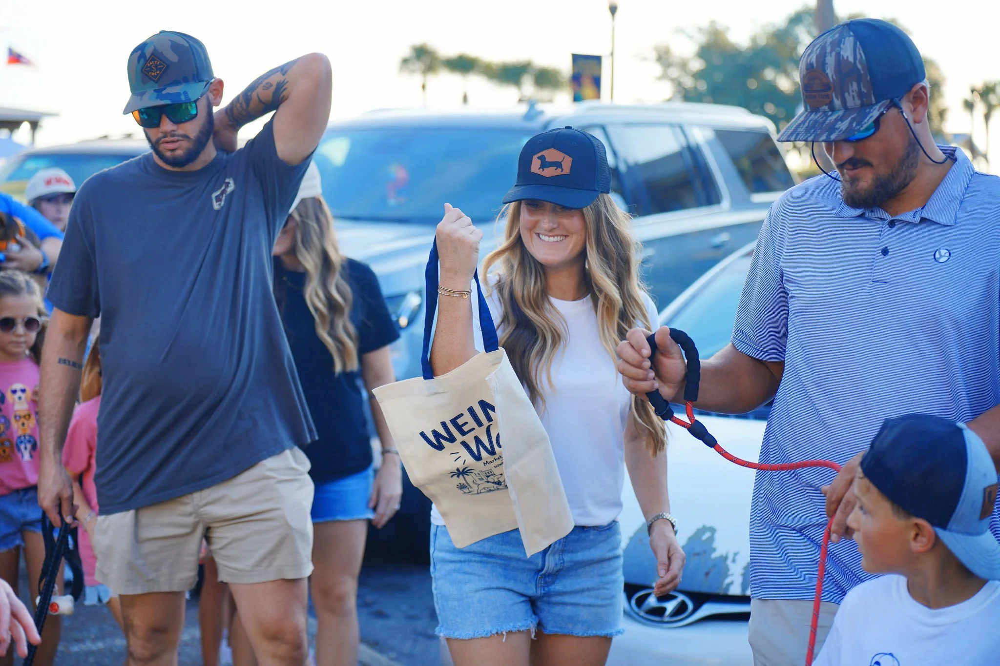 Group of people at outdoor event, some wearing hats and sunglasses, a woman smiling wearing a blue cap and a man holding a leash, with a car and trees in the background.