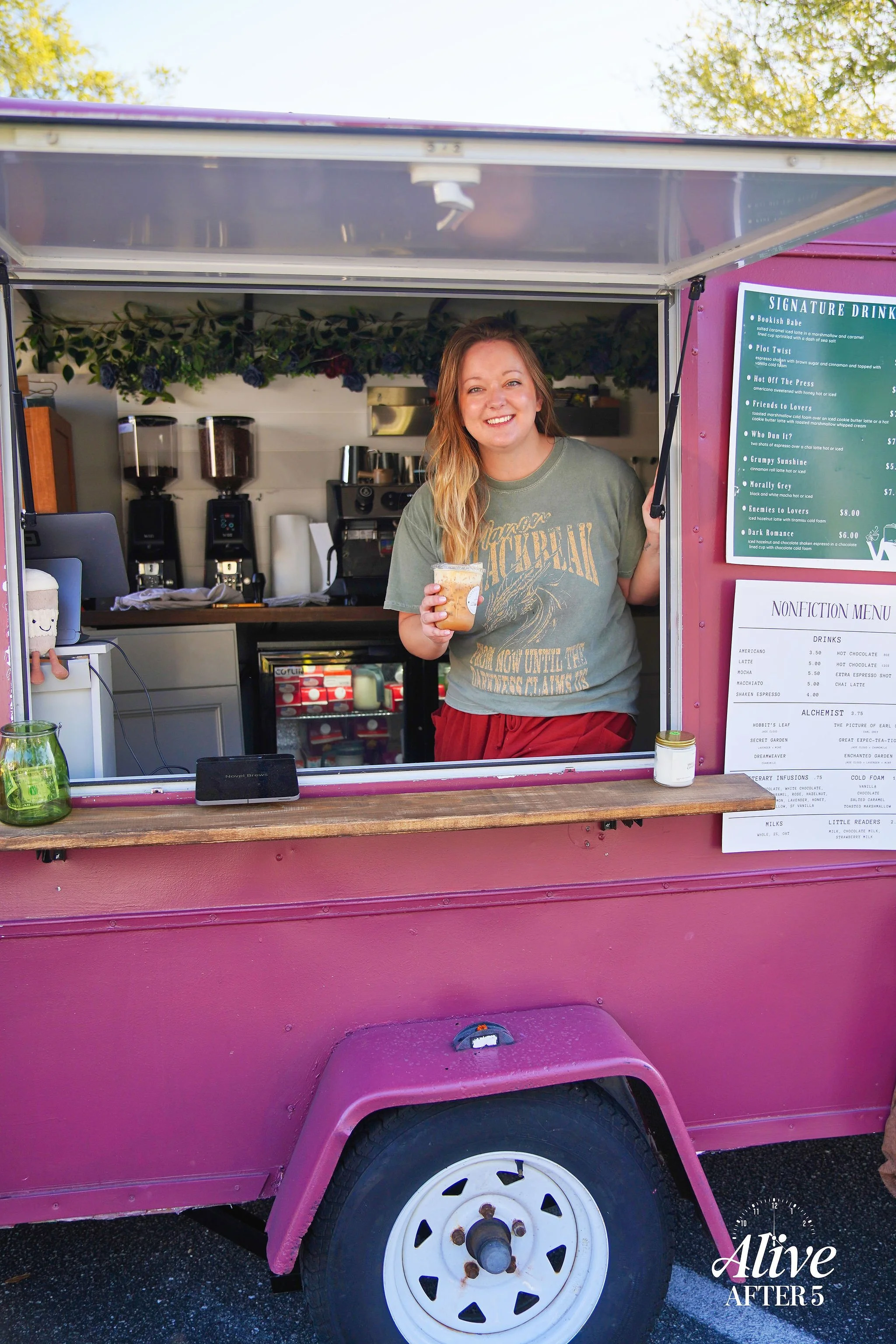 A woman with long hair and a grey T-shirt standing in a pink food truck, holding a cup, smiling at the camera. The food truck is set up outdoors with a clear sky and trees in the background.