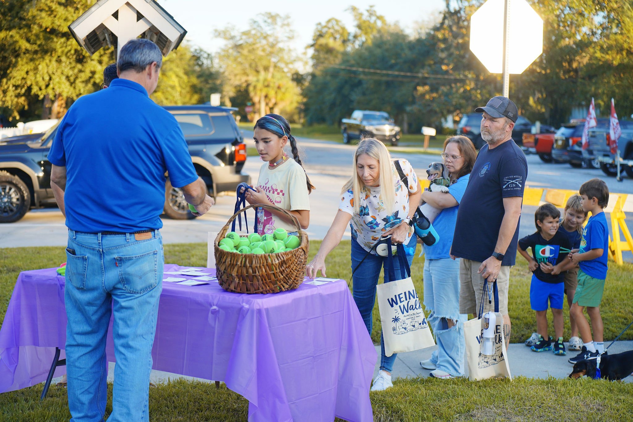 A group of people at an outdoor event near a table with a basket of tennis balls, interacting with a man in a blue shirt. The scene is set in a park in the late afternoon, with trees, cars, and a stop sign in the background.