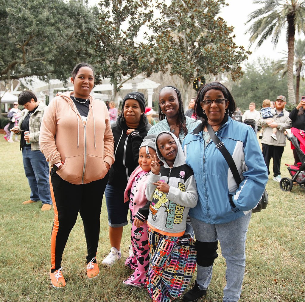 group of six people, including children and adults, standing outdoors on grass with trees and other people in the background, smiling at the camera during an outdoor event.