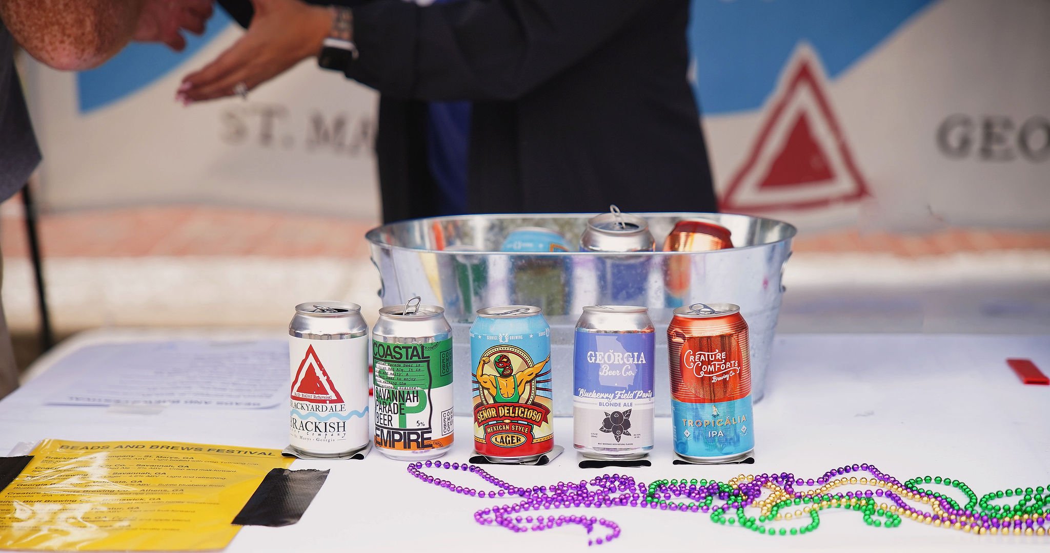 Five cans of craft beer on a white table, with colorful beads and a yellow flyer in the foreground. A man is partially visible behind the cans, reaching into a metal bucket filled with more cans. The background shows a banner with the words 'St. Ma' 