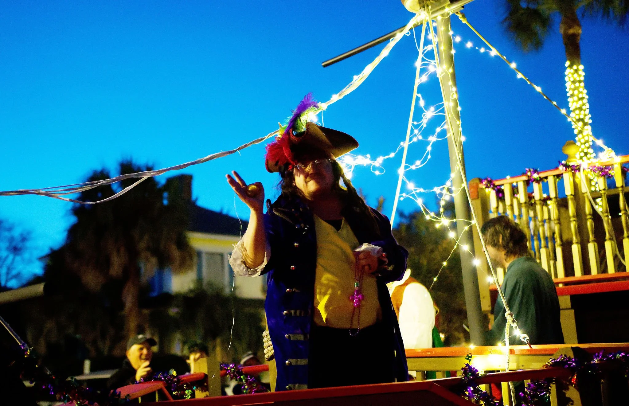 Person dressed as a pirate wearing a colorful feathered hat, standing on a decorated float with string lights during a nighttime parade or celebration.