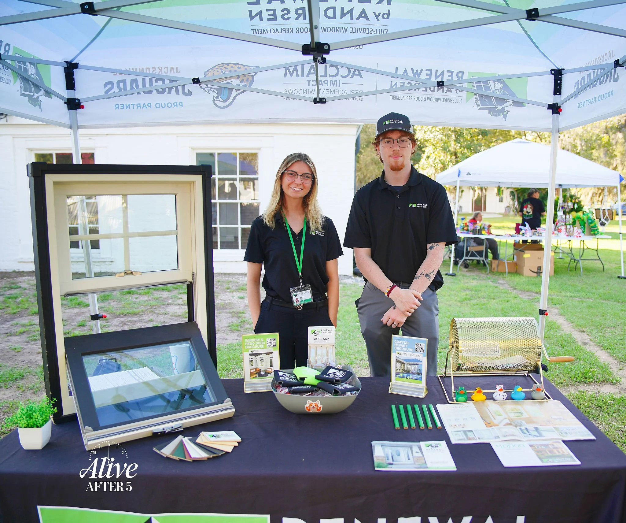 Two people standing behind a table at an outdoor booth, smiling, with pamphlets, a window display, and small rubber duck toys on the table.