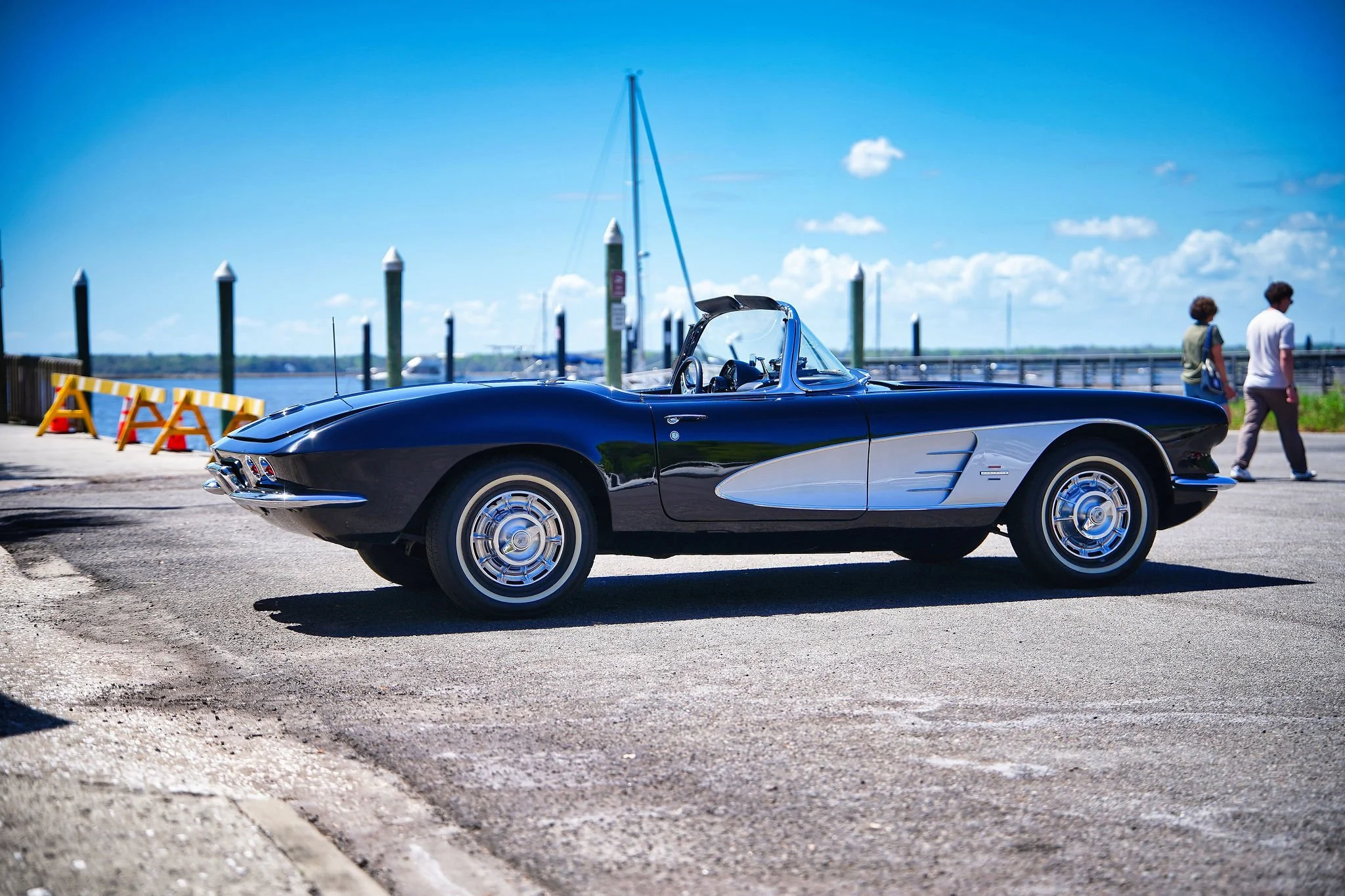 A vintage black and silver Corvette convertible parked near a waterfront with sailboats in the background, two people walking nearby on a sunny day.