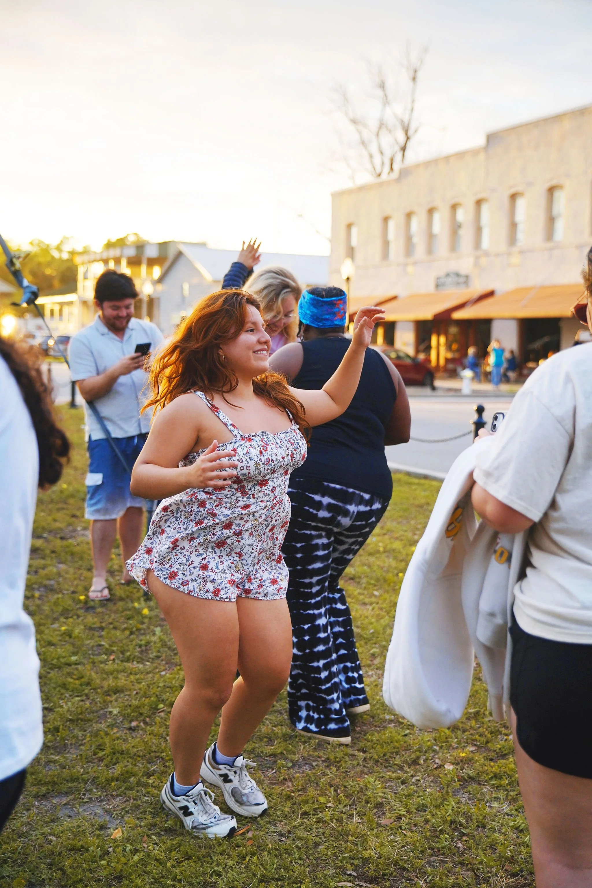 A group of diverse people dancing outdoors on a grassy area during sunset, with buildings and a street in the background.