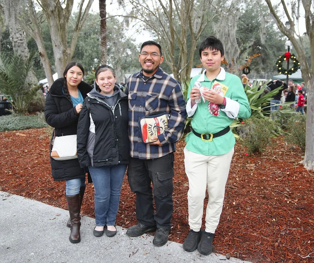 Group of four people standing outdoors during a holiday event, with Christmas wreaths and festive decorations in the background.