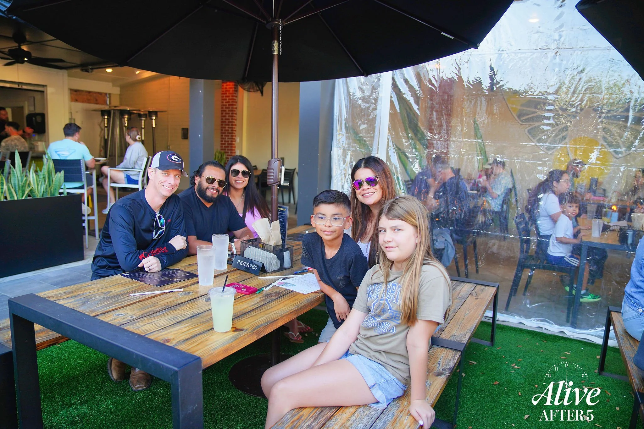 Group of six people sitting at an outdoor restaurant table, smiling, with drinks and food, under a large patio umbrella, with a clear plastic view of other diners in the background.