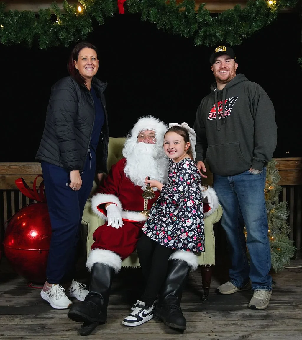 Family photo with Santa Claus at Christmas, with a woman, a girl holding a bell, and a man, standing beside Santa seated on a chair, in front of a Christmas tree decorated with lights and ornaments.