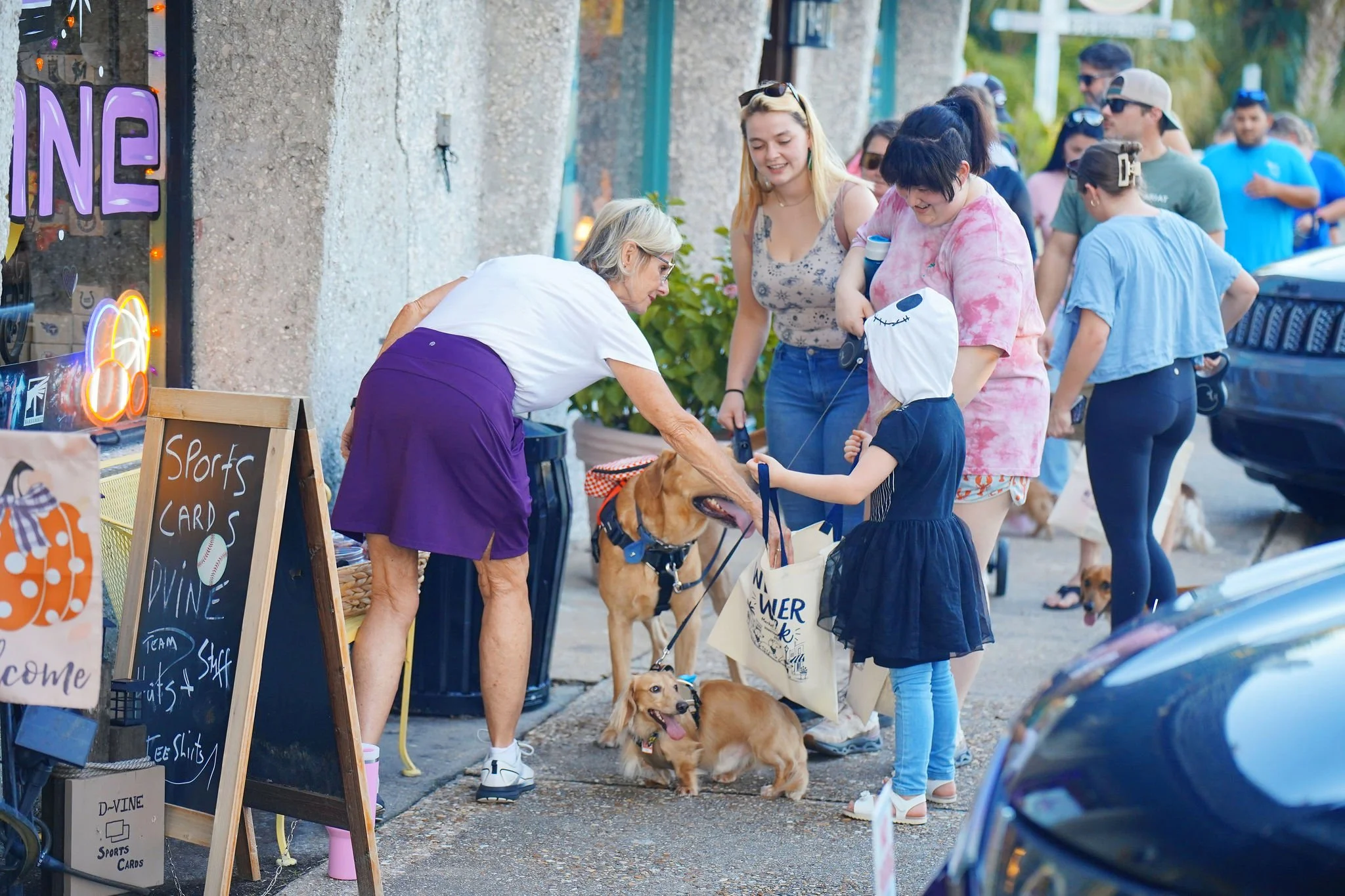 People gathered outside a storefront with dogs, shopping bags, and signs selling sports cards. Some individuals are interacting with the dogs at a pet-friendly event on a sidewalk.
