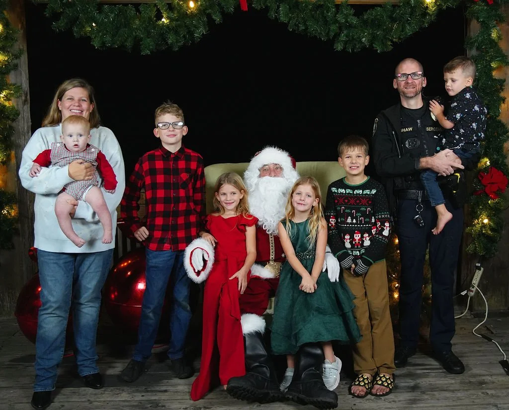 Family posing with Santa Claus for Christmas photo, surrounded by holiday decorations including large red ornaments and greenery.