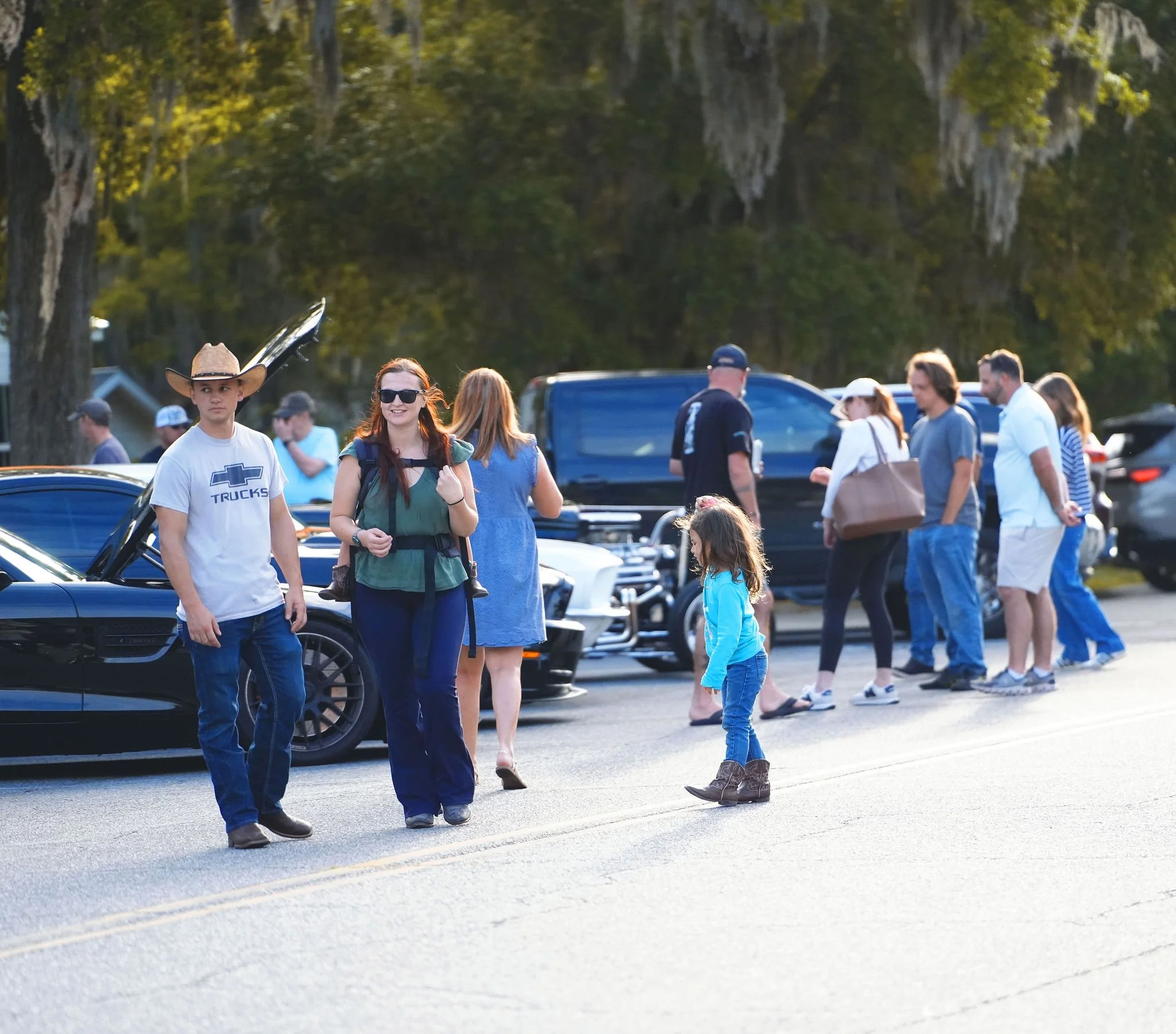 People walking on the street, some with vintage cars parked along the side, trees in the background, during daylight.