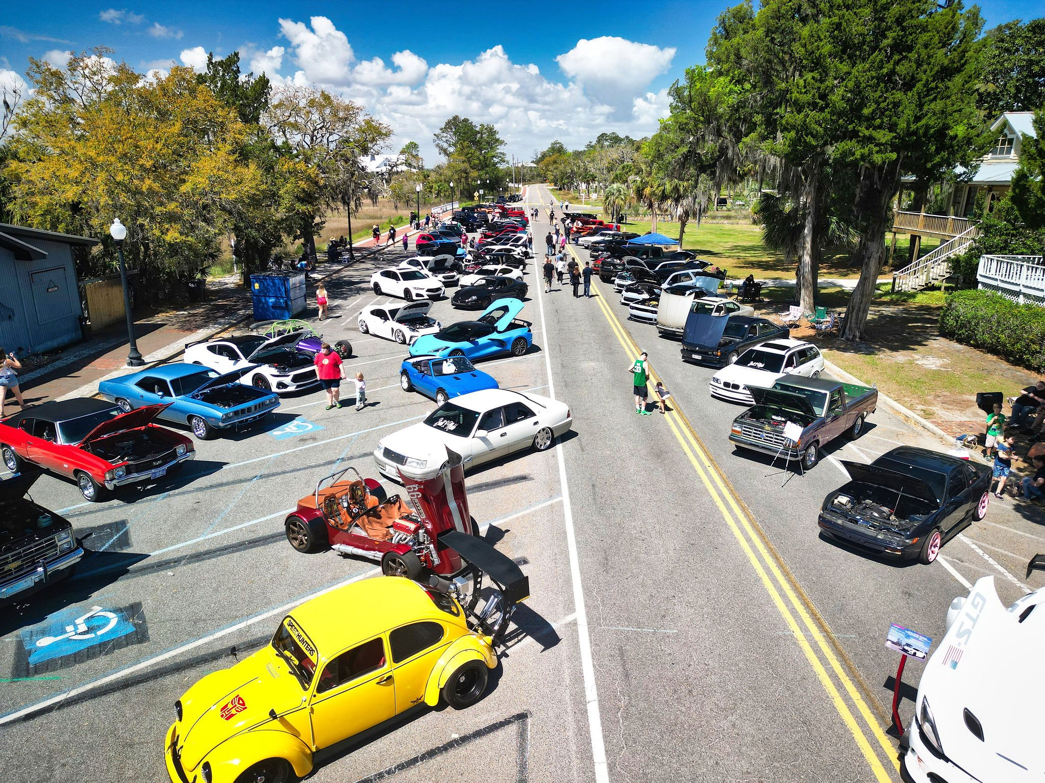 A car show with various cars on display along a street, including classic, sports, and modified vehicles, with people walking around and viewing the cars on a sunny day with partly cloudy skies.