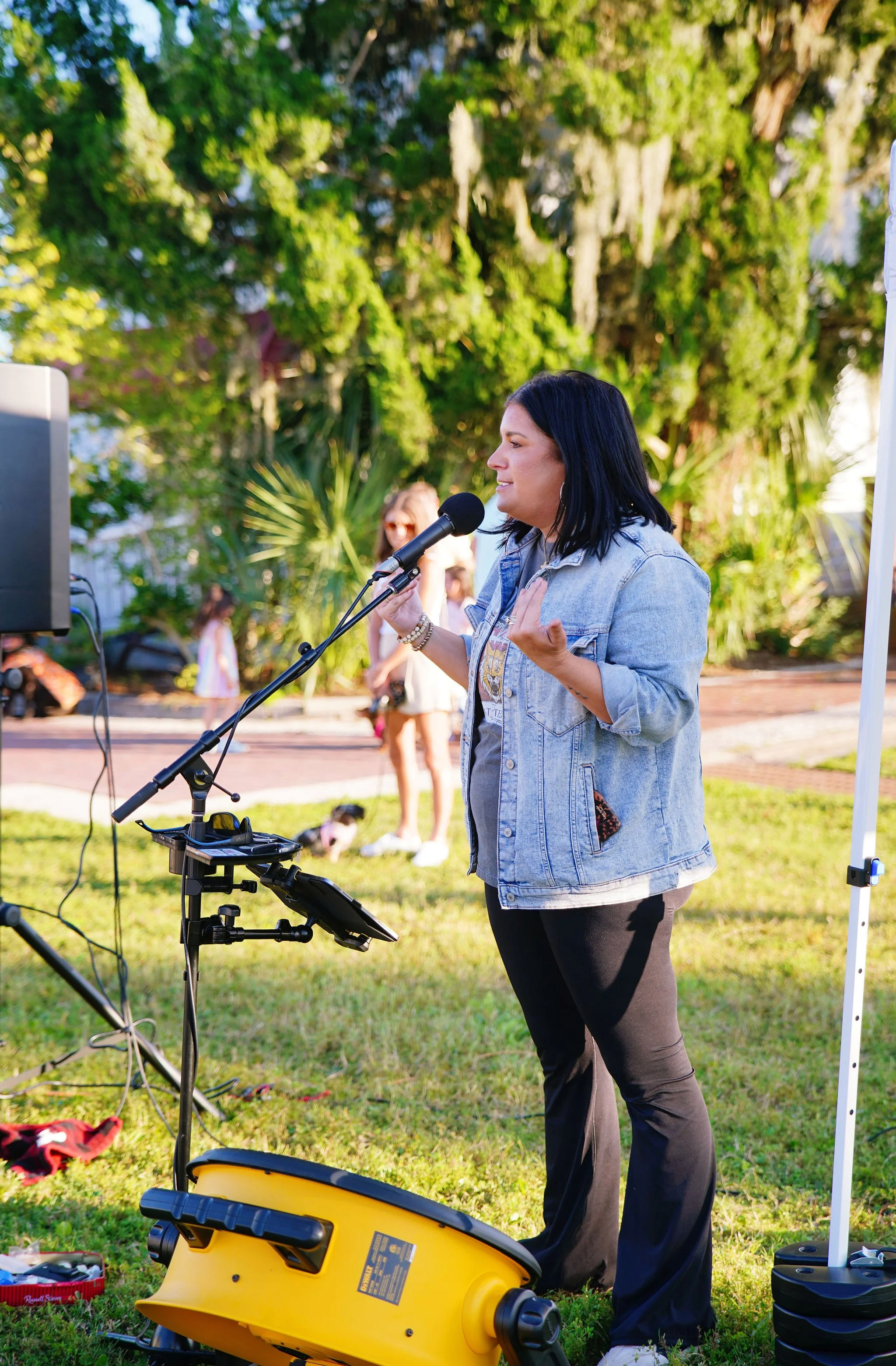 A woman with dark hair speaking into a microphone outdoors, with green trees and people in the background, next to a yellow extension cord reel and equipment.